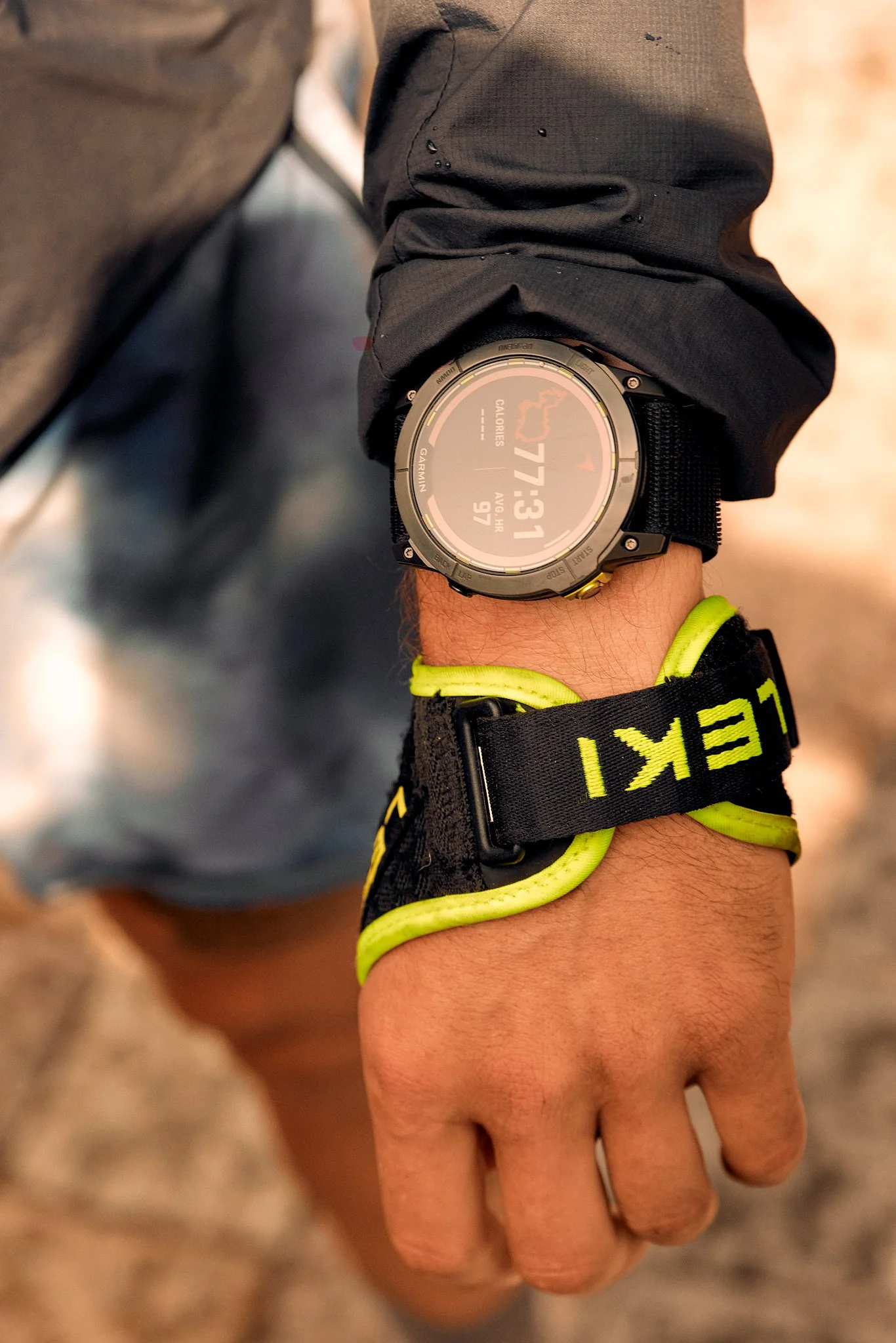 A person wearing a black and yellow LEKI wrist strap and a black watch with a digital display showing calories burned, date, and time, and a black/gray jacket with water droplets on the sleeve, with a blurred background.