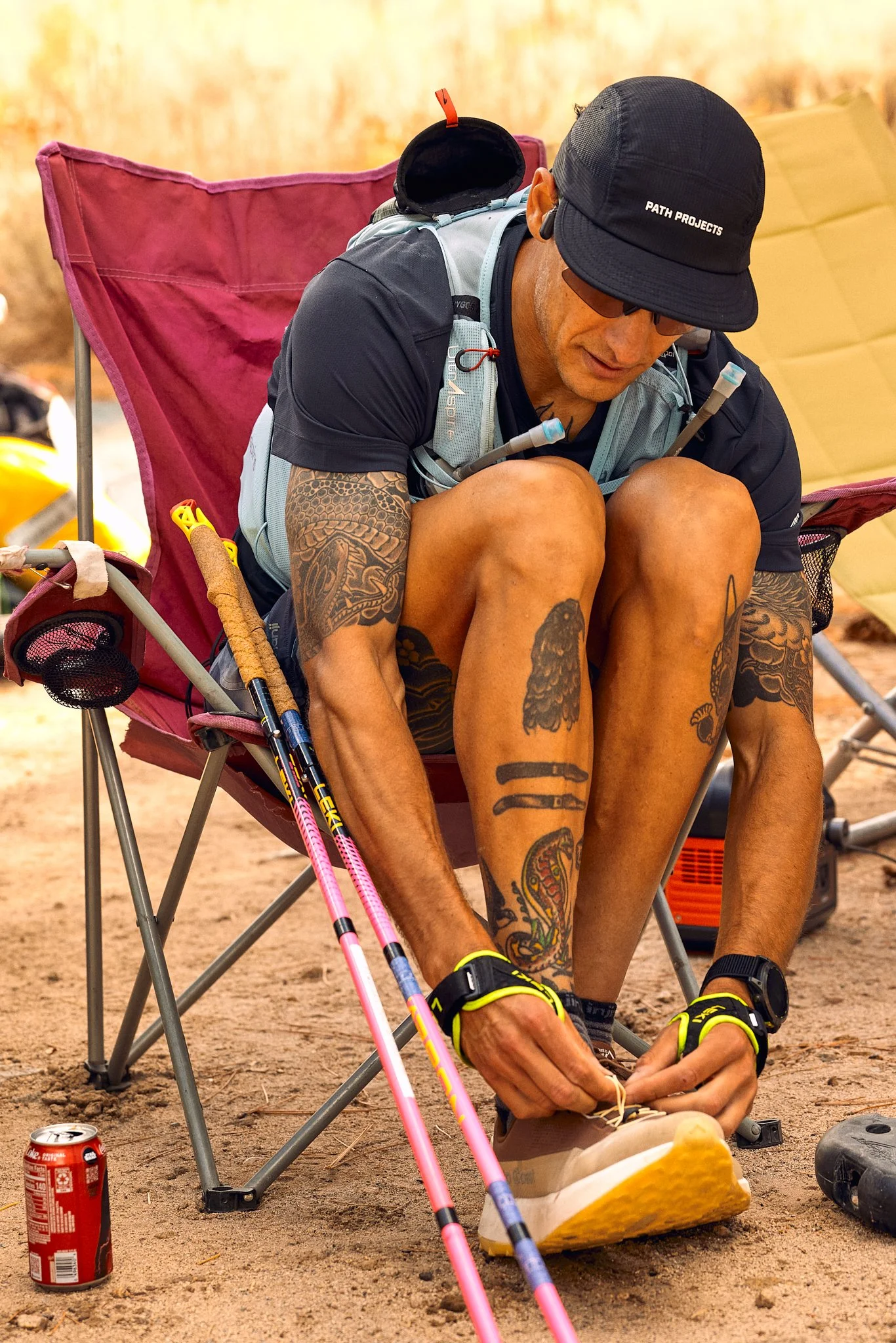 A man with tattoos, wearing a black cap with 'PATH PROJECTS' written on it, sitting in a portable chair outdoors, tying a shoe. He has hiking poles and a backpack, with a goldfish-shaped foam or pillow on his lap, a can of Coca-Cola on the ground, an