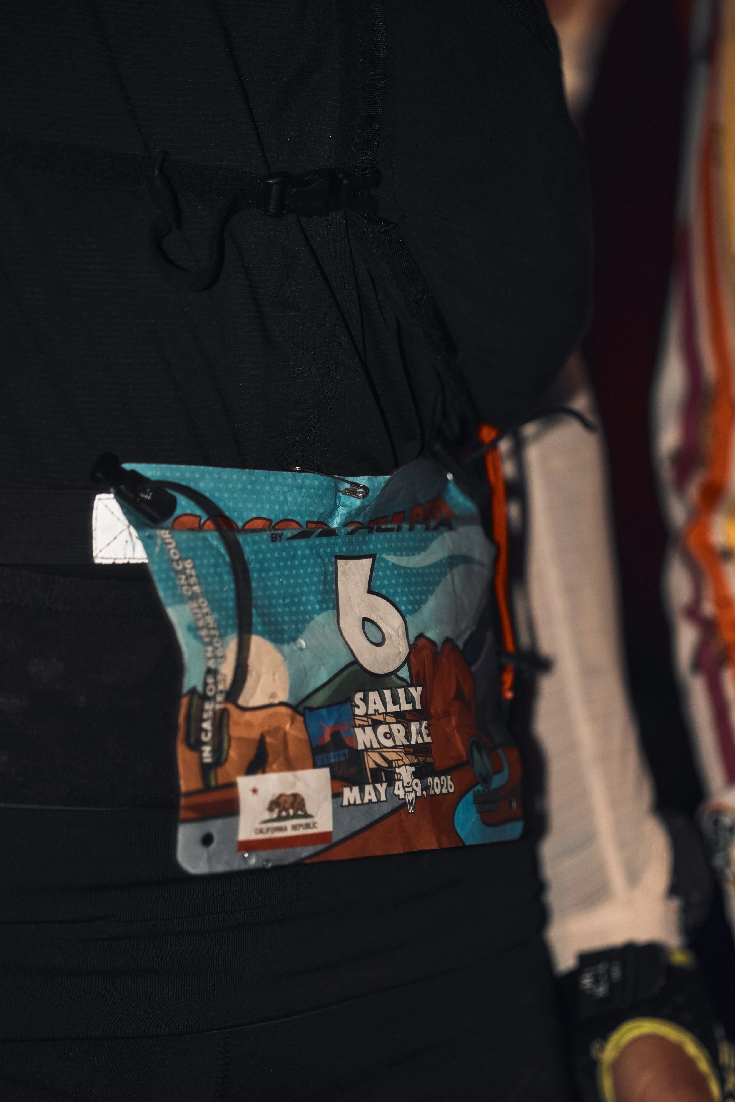 Close-up of a race bib with the name Sally Morgan, featuring colorful graphics and a bear, attached to a person's black athletic clothing.