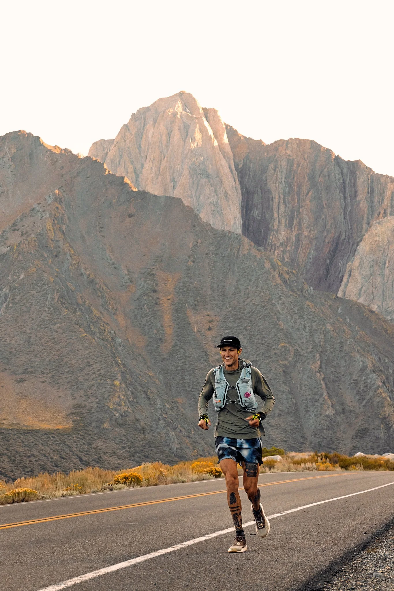 A male trail runner with a backpack running on a mountain road with a mountain range in the background during golden hour.