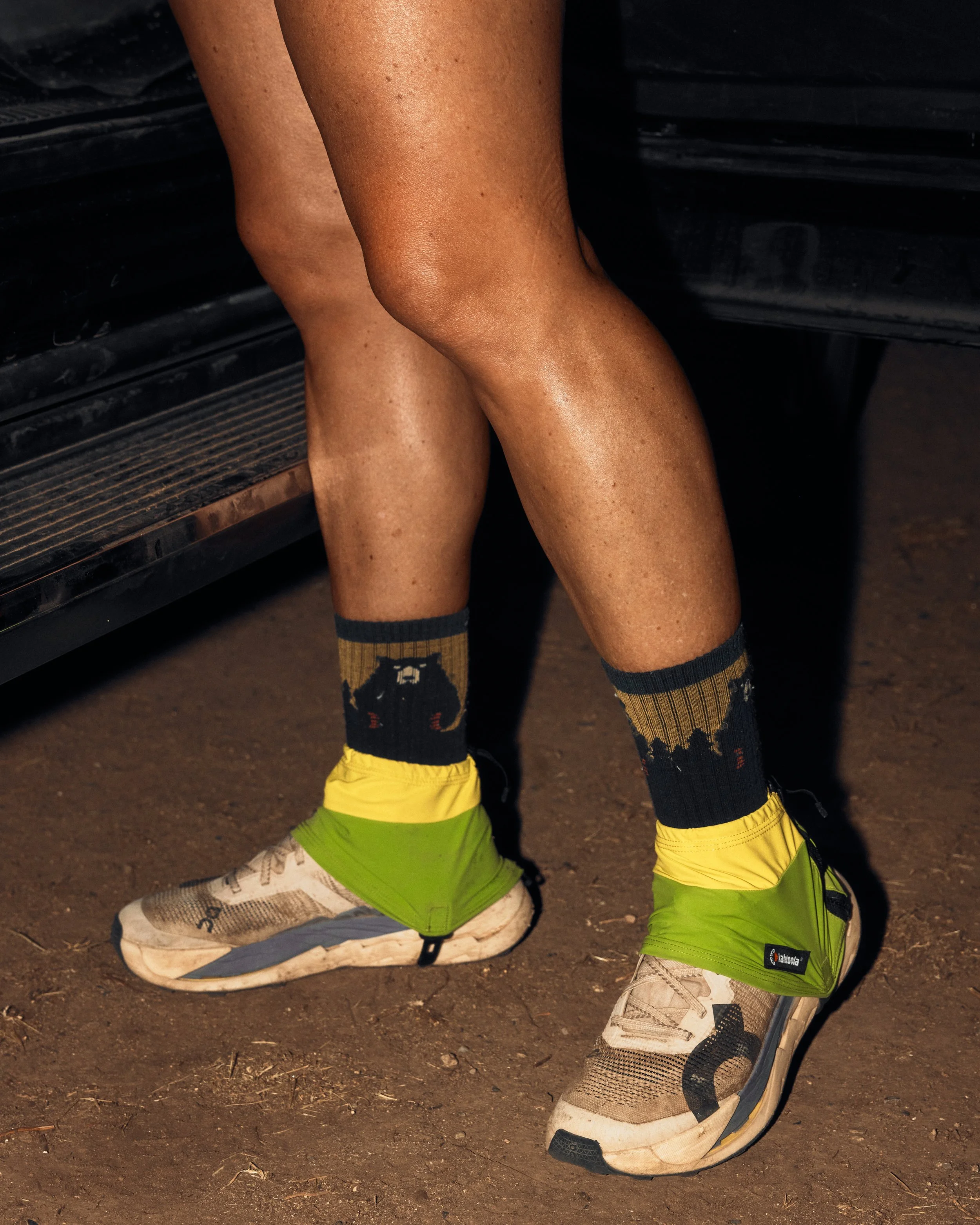Close-up of person's legs wearing bright yellow shoes, beige and black "ON"running shoes, green compression shorts with black logos, black and yellow patterned socks, and standing on dirt ground.