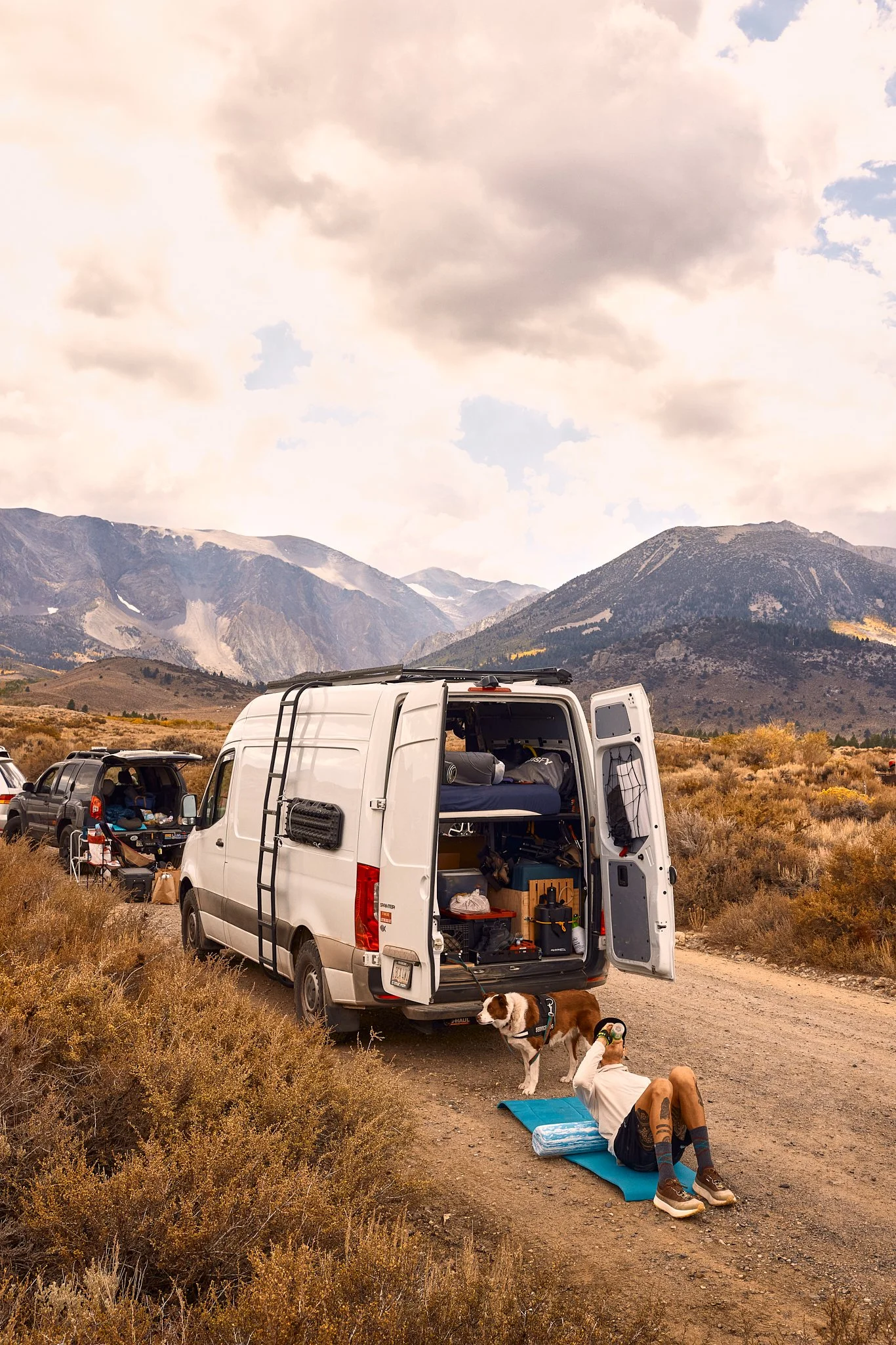An outdoor scene with a white camper van and several motorcycles parked on a dirt road in a mountainous area. A person is lying on a yoga mat doing a stretch, with a dog nearby. The background features mountains under a cloudy sky, and supplies are s