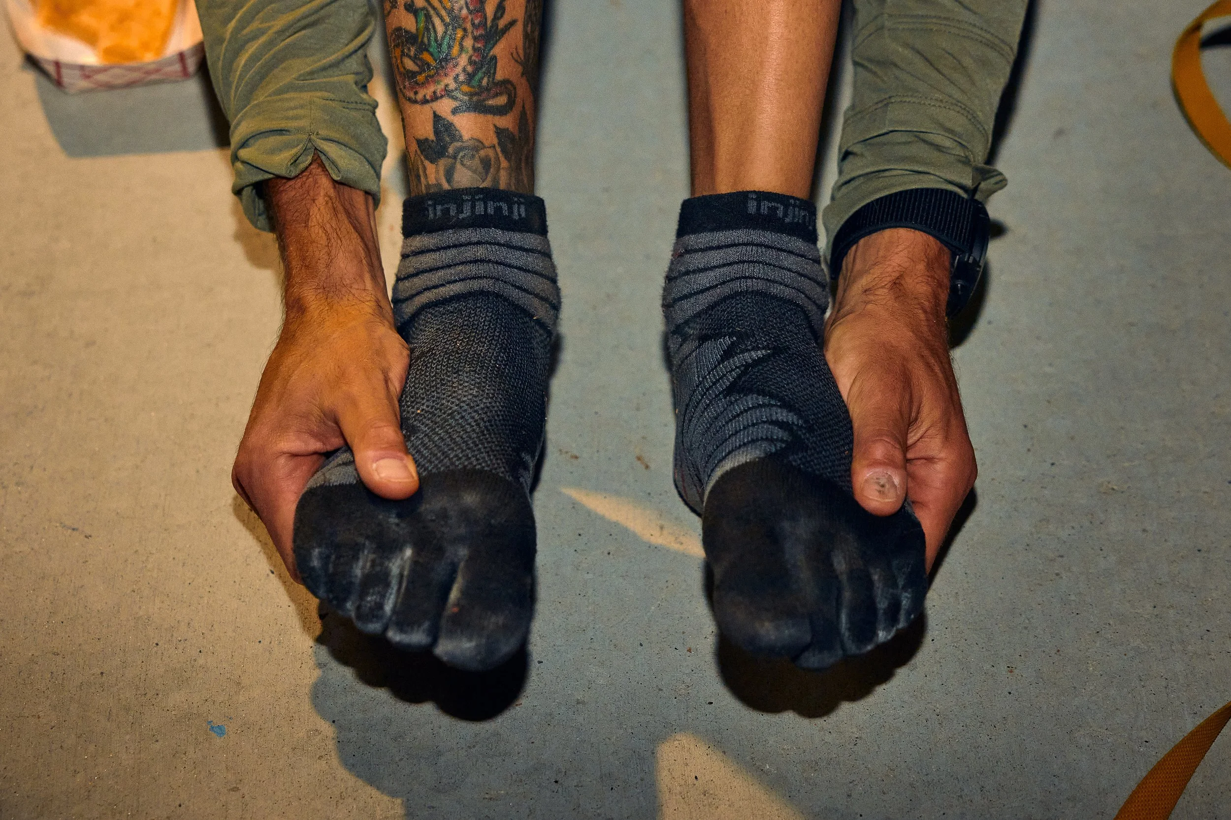Person with tattoos on their left arm wearing dark socks and black shoes, seated on a concrete floor, with their legs extended, feet pointing outward, and hands resting on their ankles.