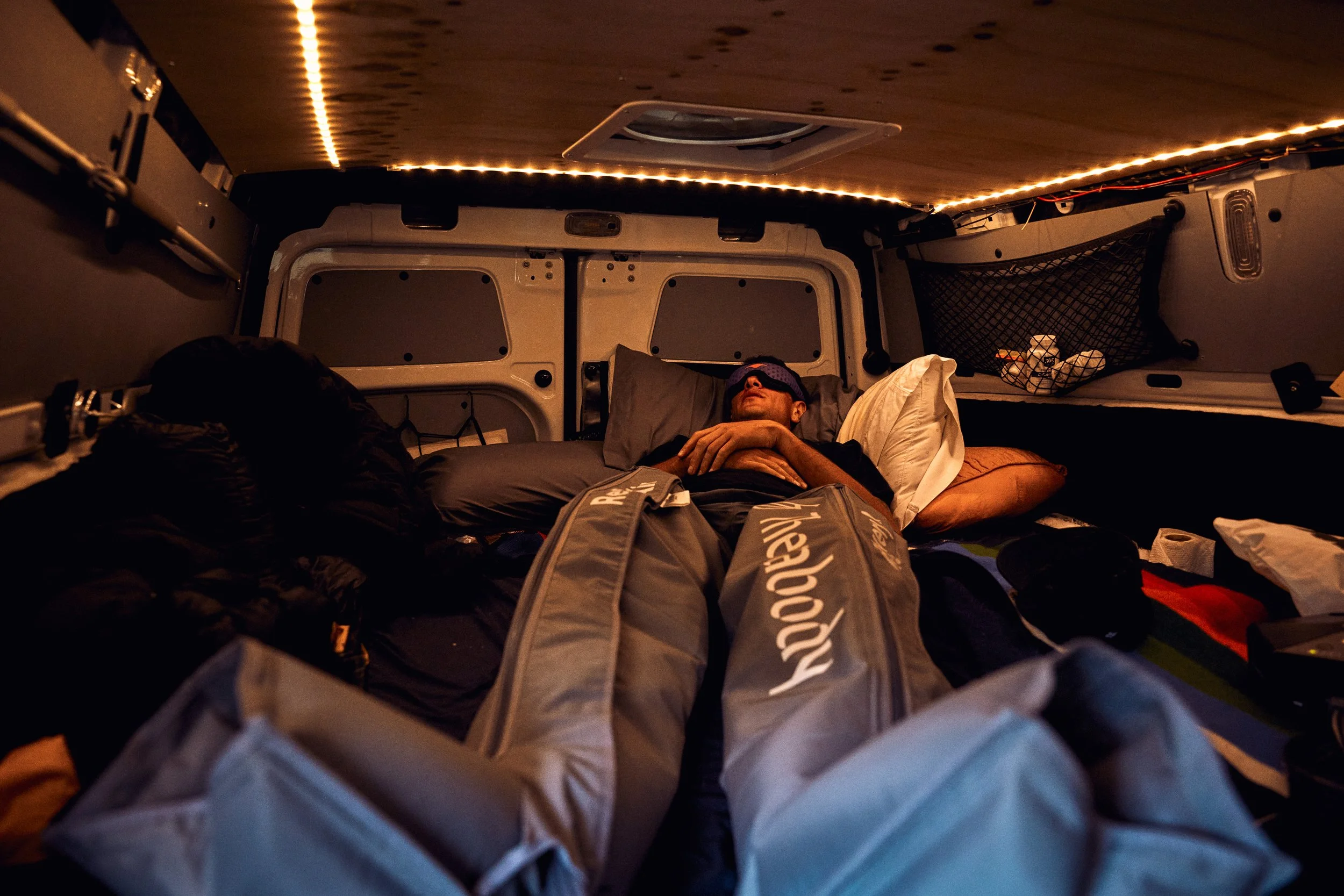 Inside of a van or small camper with a person resting on a bed, wearing an eye mask. The ceiling is lit with LED strip lights, and there are various belongings and storage nets around.