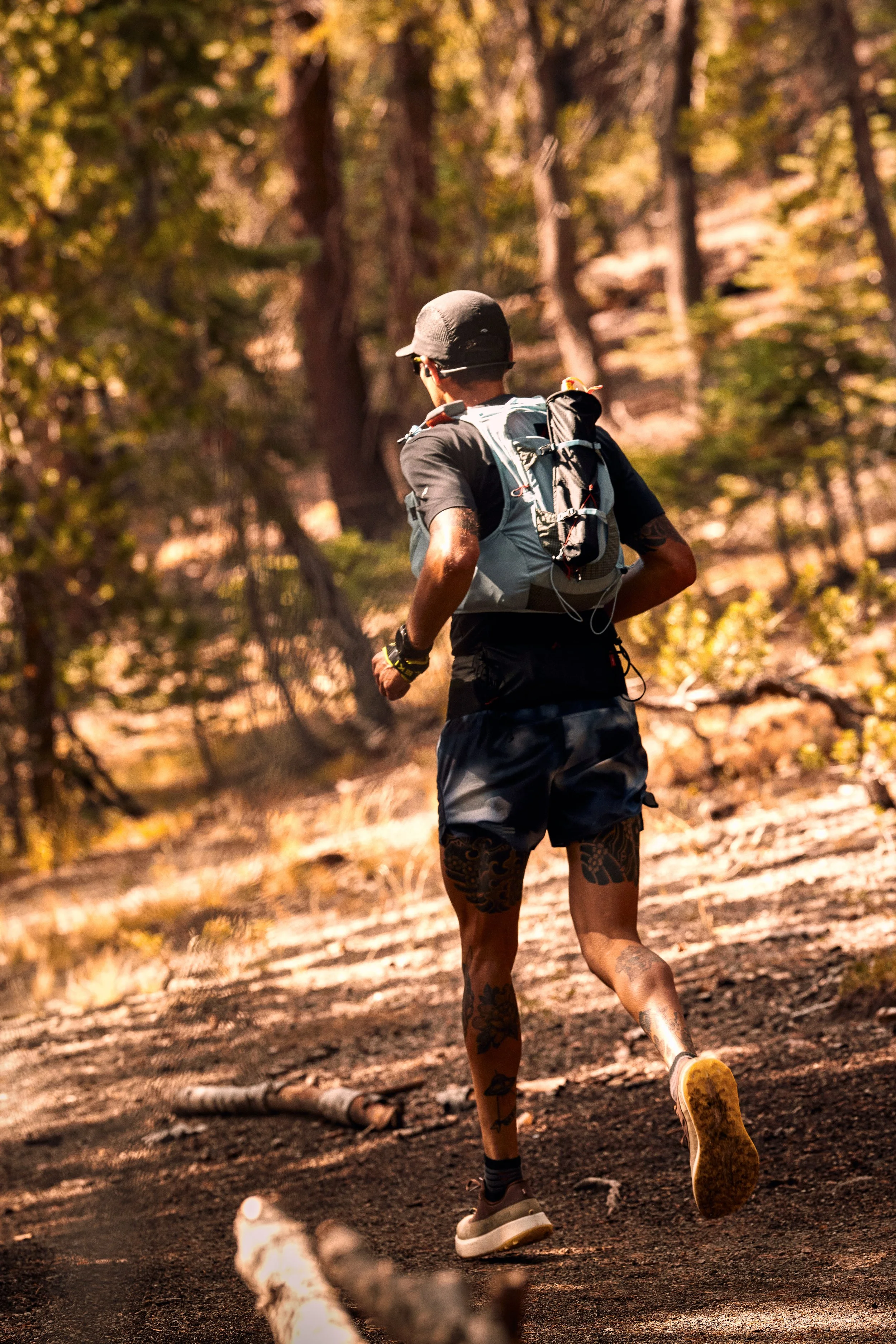 A person running on a dirt trail through a forest with tall trees and autumn leaves, wearing a gray hat, black shirt, and shorts with tattoos on the legs.