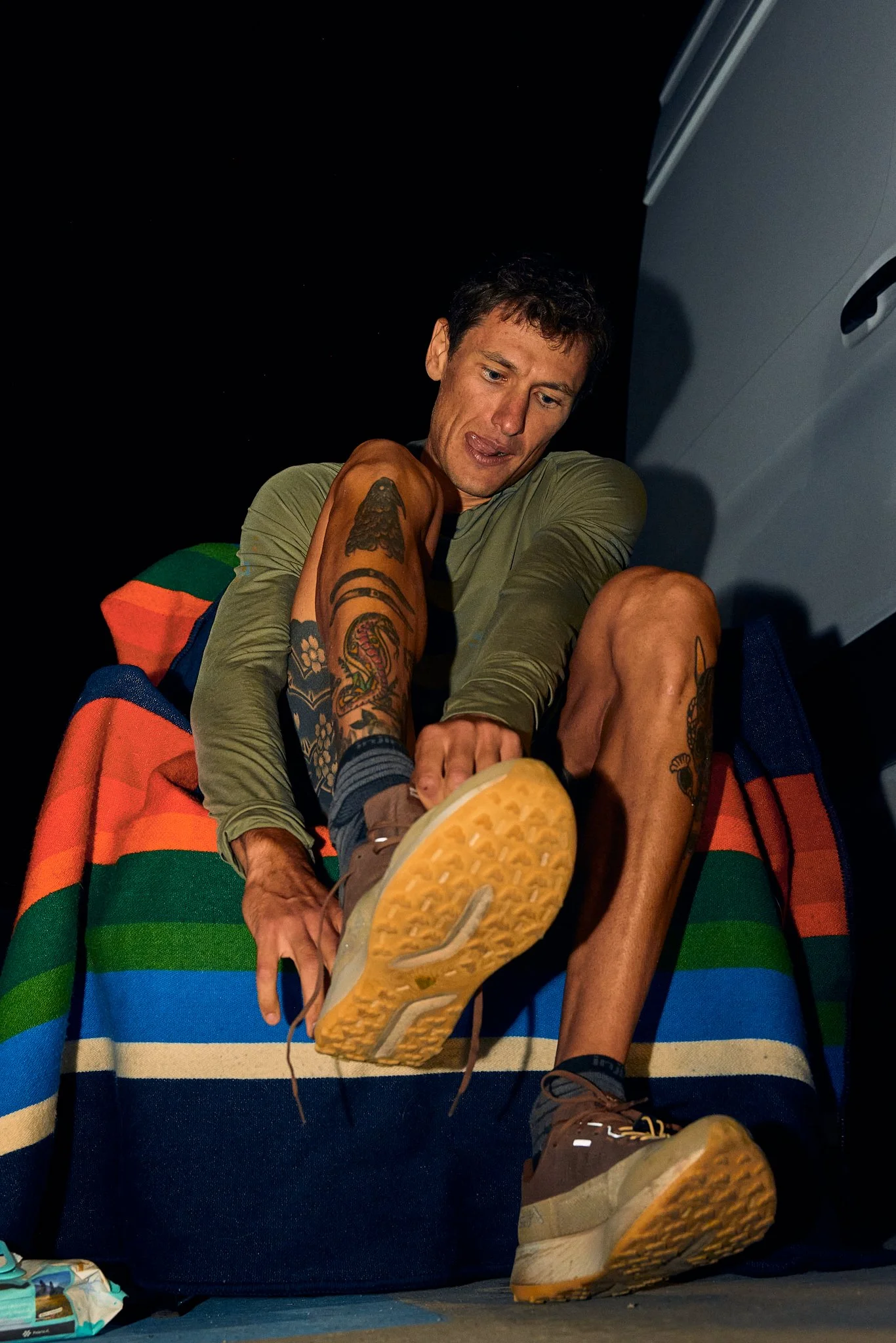 A man with tattoos on his arms sitting on a colorful striped blanket at night, wearing hiking shoes, with his tongue slightly out, appearing to be in a playful or relaxed mood.
