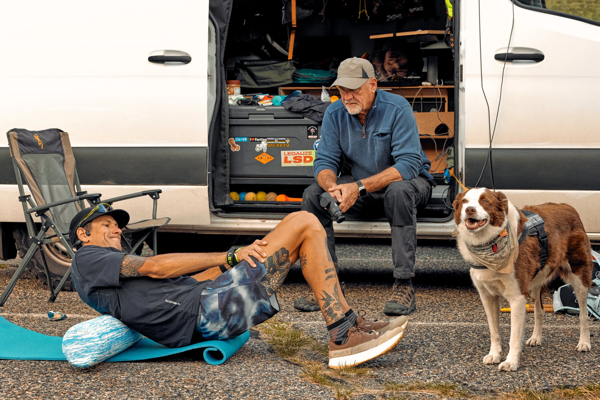 Two men and a dog outdoors next to a white van. One man with tattoos lying on a mat, the other man sitting in front of the van's open storage area. A dog wearing a bandana is standing nearby. The van's interior has various supplies and stickers, incl