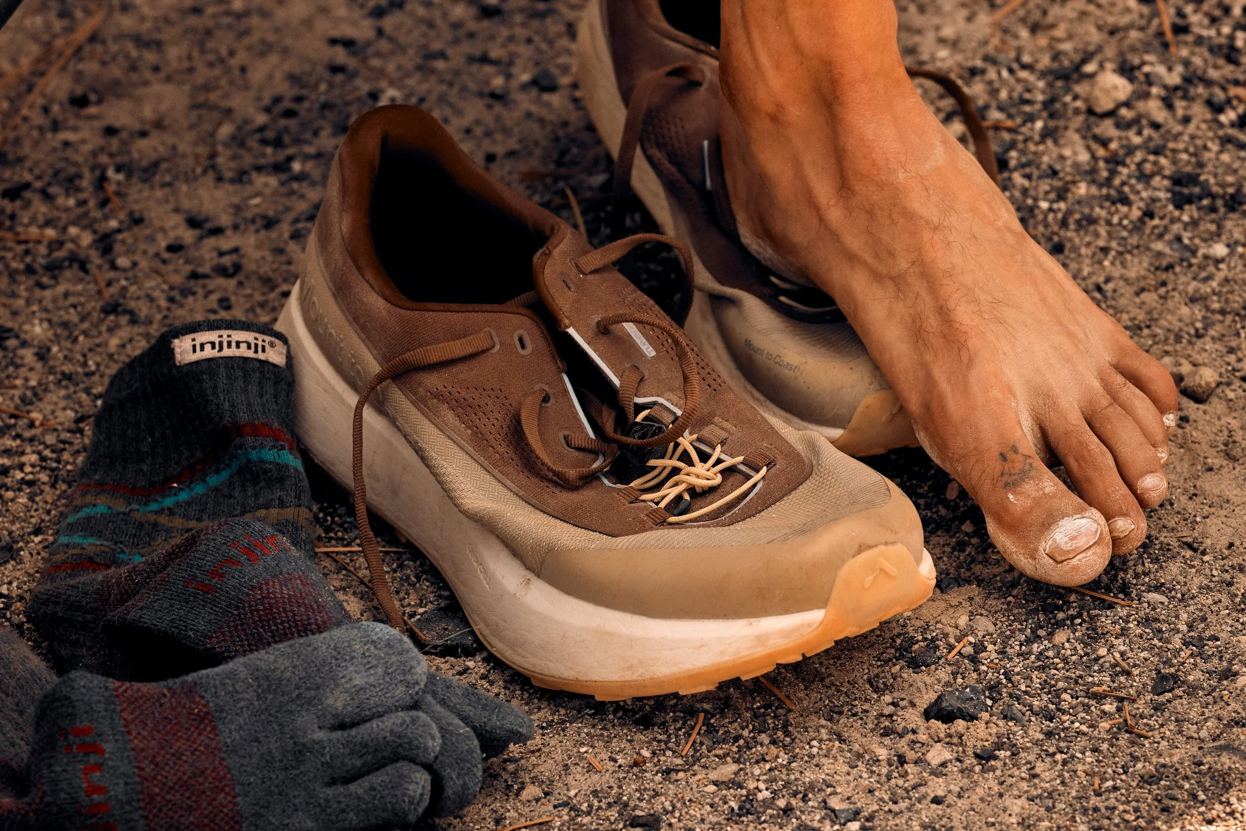 A person's foot is placed on the ground next to a beige sneaker with brown accents, surrounded by dirt and small rocks. The person has a tattoo on their toes and visible hair on their foot. A sock with red and teal stripes is partially visible in the