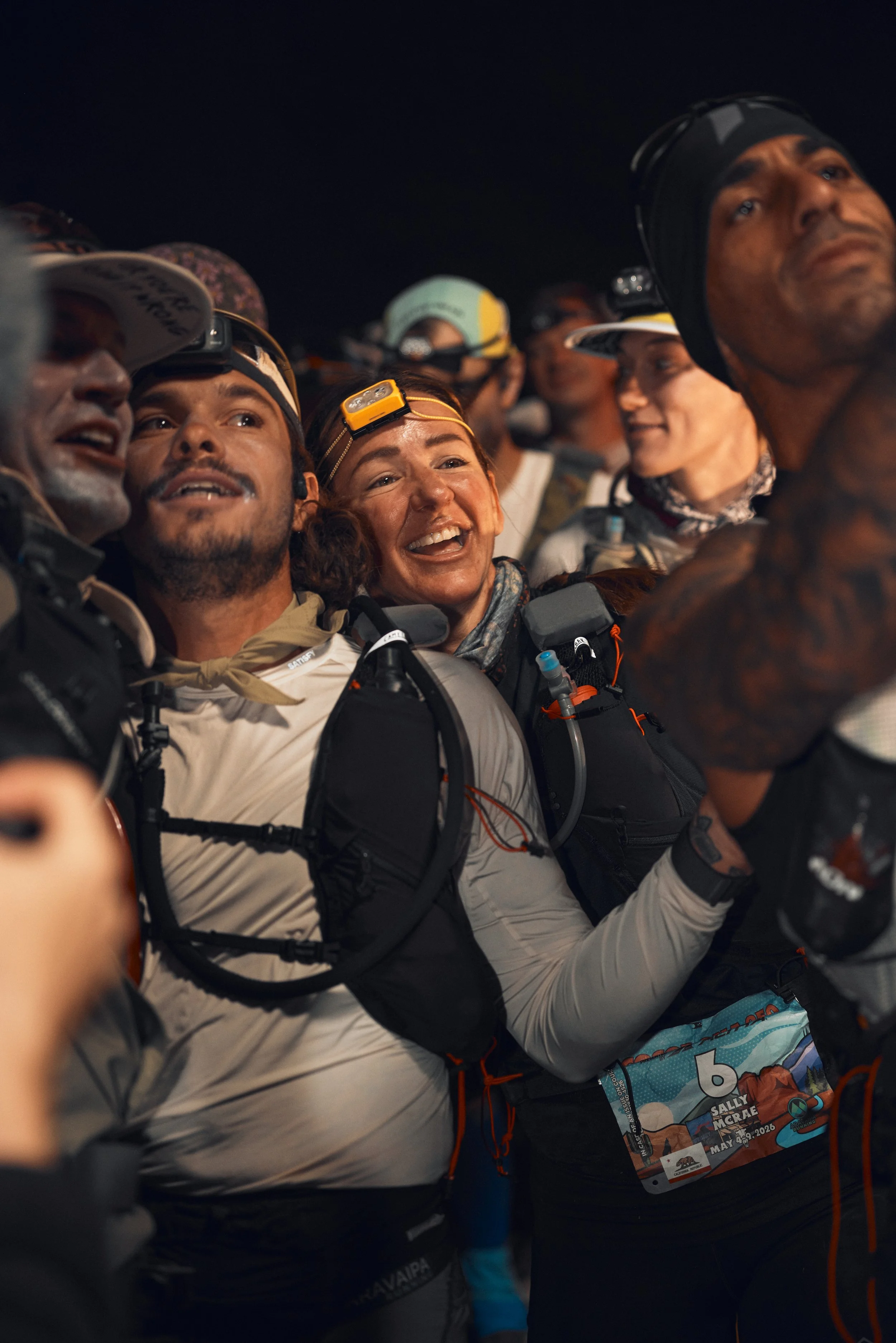 Group of people wearing headlamps and athletic gear, smiling and gathered closely at night, Cocodona 250
