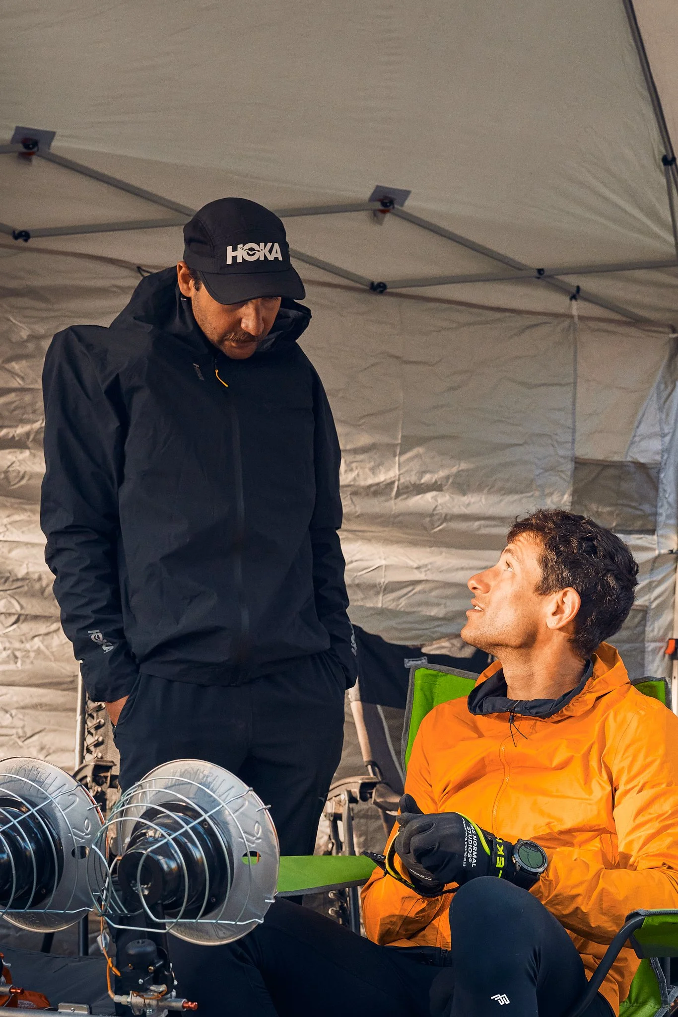 Two men in outdoor gear, one standing and one sitting, under a tent with equipment and fans, engaging in conversation.