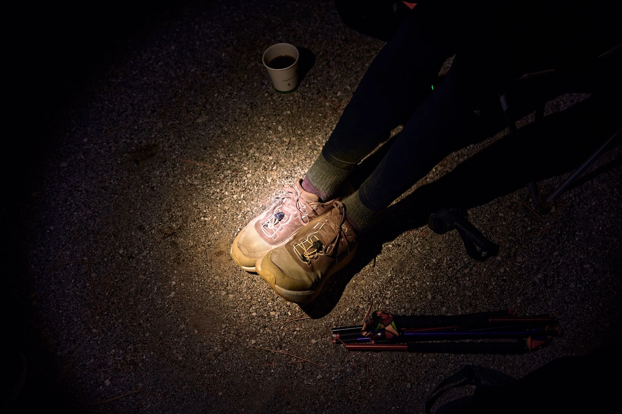 Close-up of a person's legs and feet wearing hiking shoes and pants, standing on a gravel surface at night. There is a coffee cup on the ground and part of a tripod or hiking pole is visible to the right.