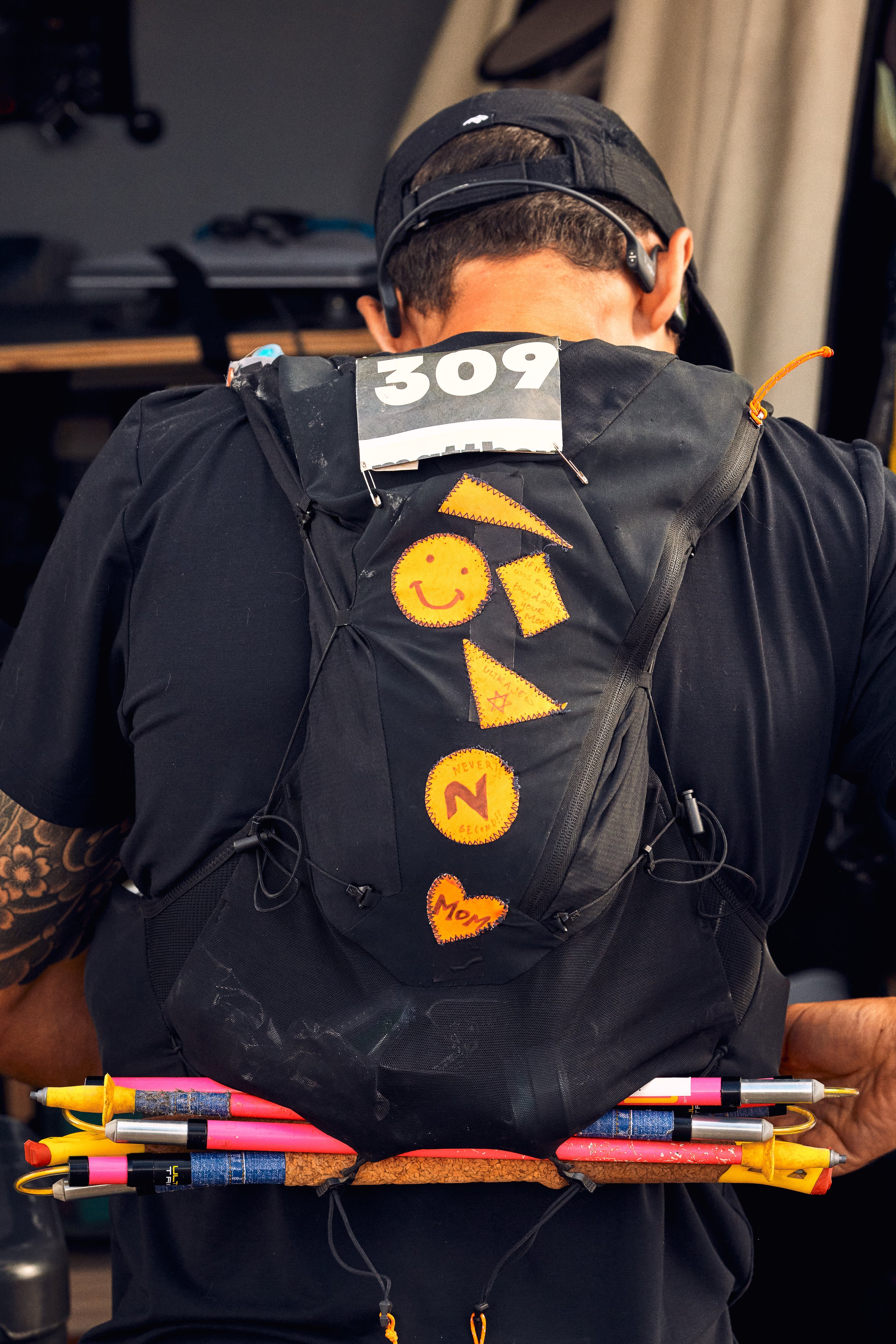 A person with a black backpack decorated with orange smiley faces and handwritten notes, wearing a black cap and headphones, holding a stack of colorful pens.