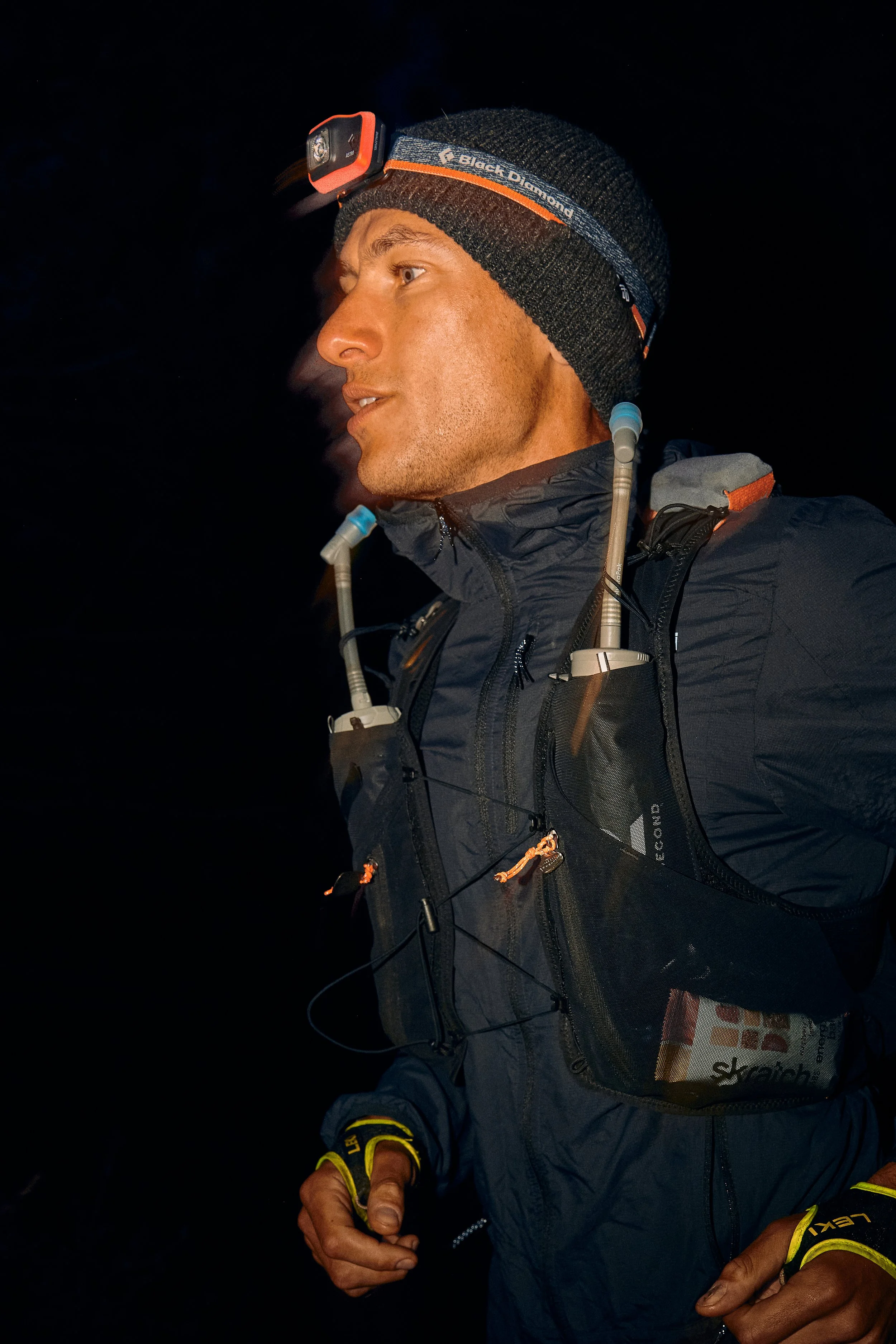 A man wearing a black outdoor jacket and a gray knit beanie with a mounted headlamp. He has a hydration tube over his shoulder and yellow and black gloves, with a dark background.