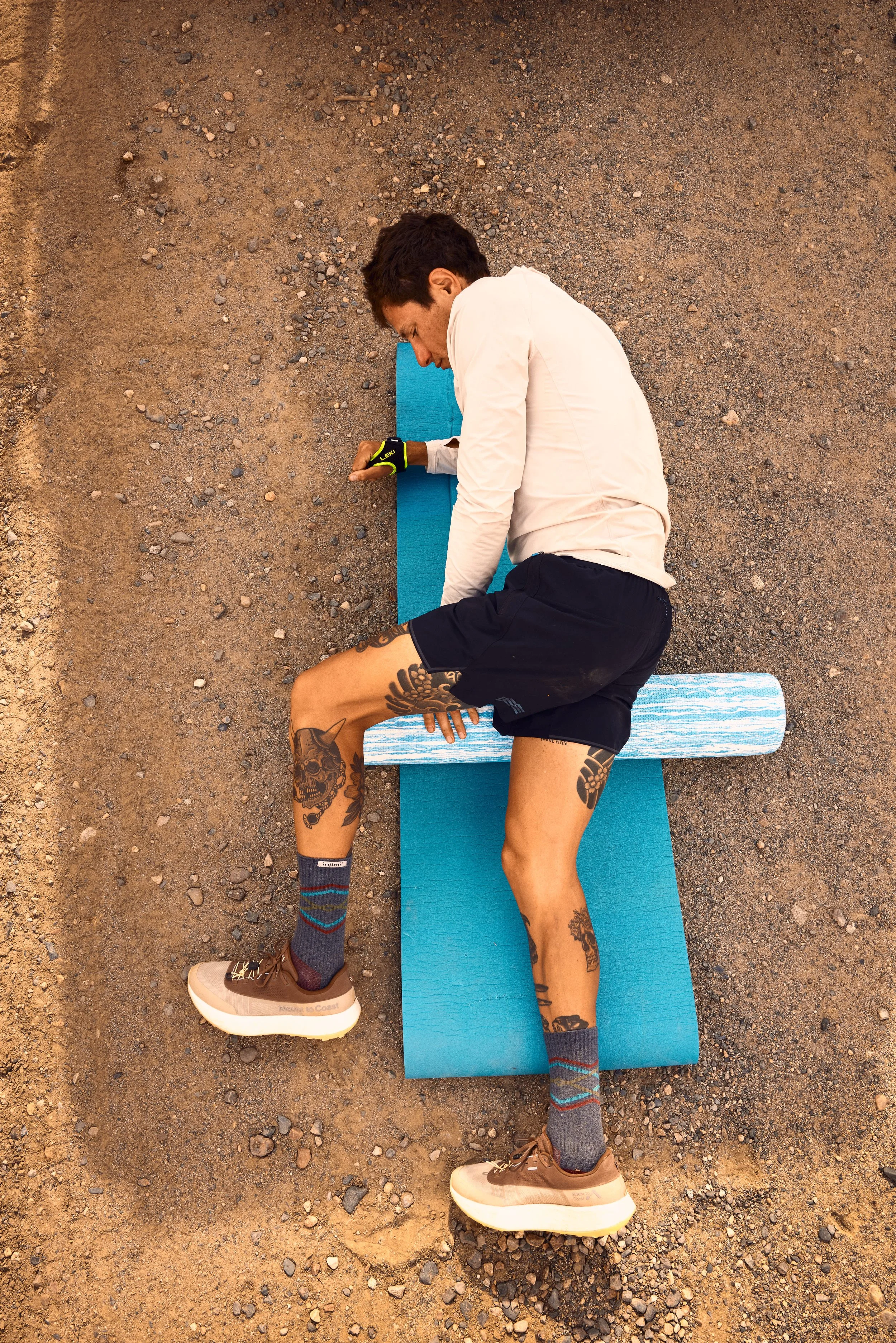 A man lying on an exercise mat outdoors on rocky ground, stretching his leg.