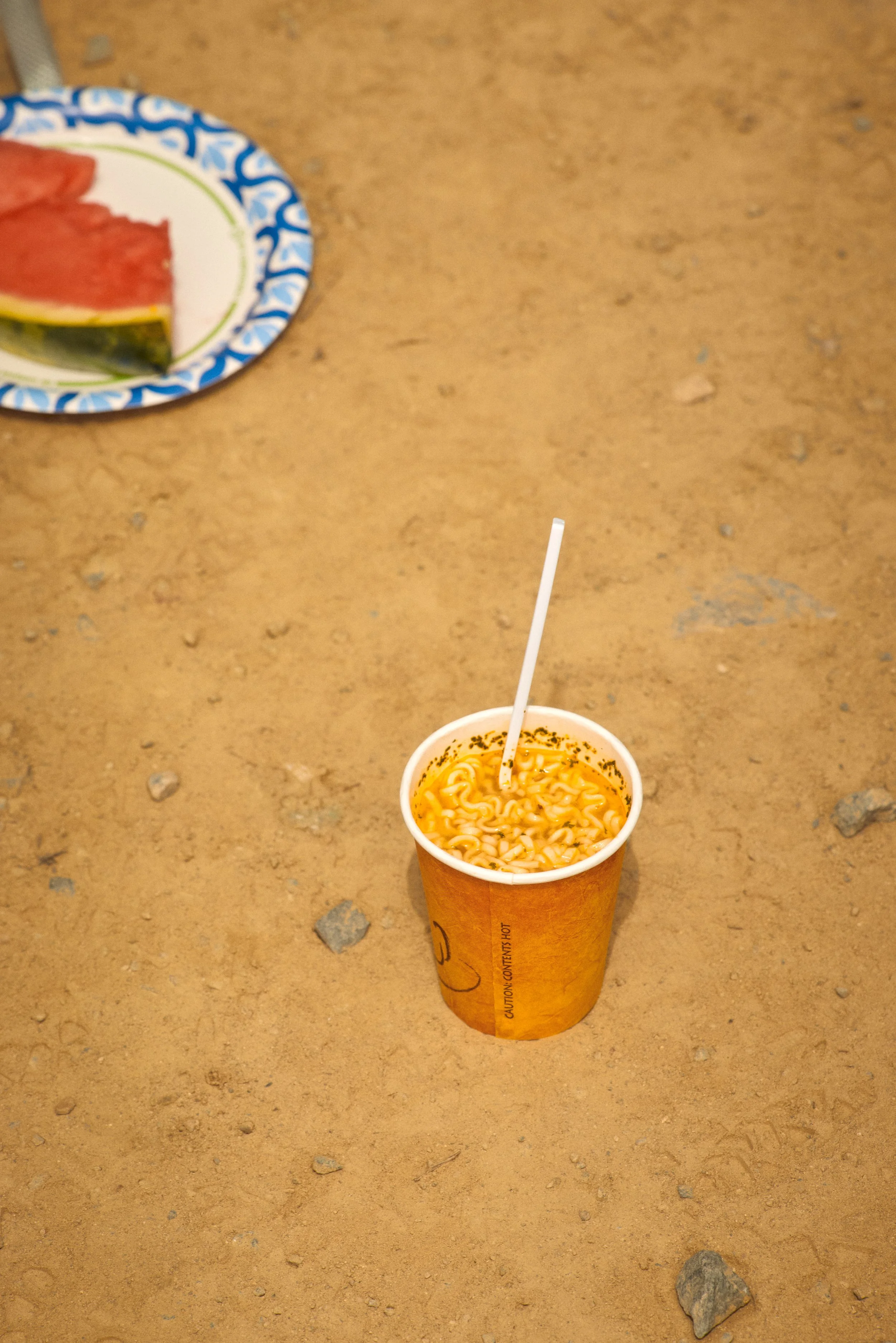 A cup of instant ramen noodles with a straw on a sandy surface, with a plate of watermelon slices on a decorative paper plate in the background.