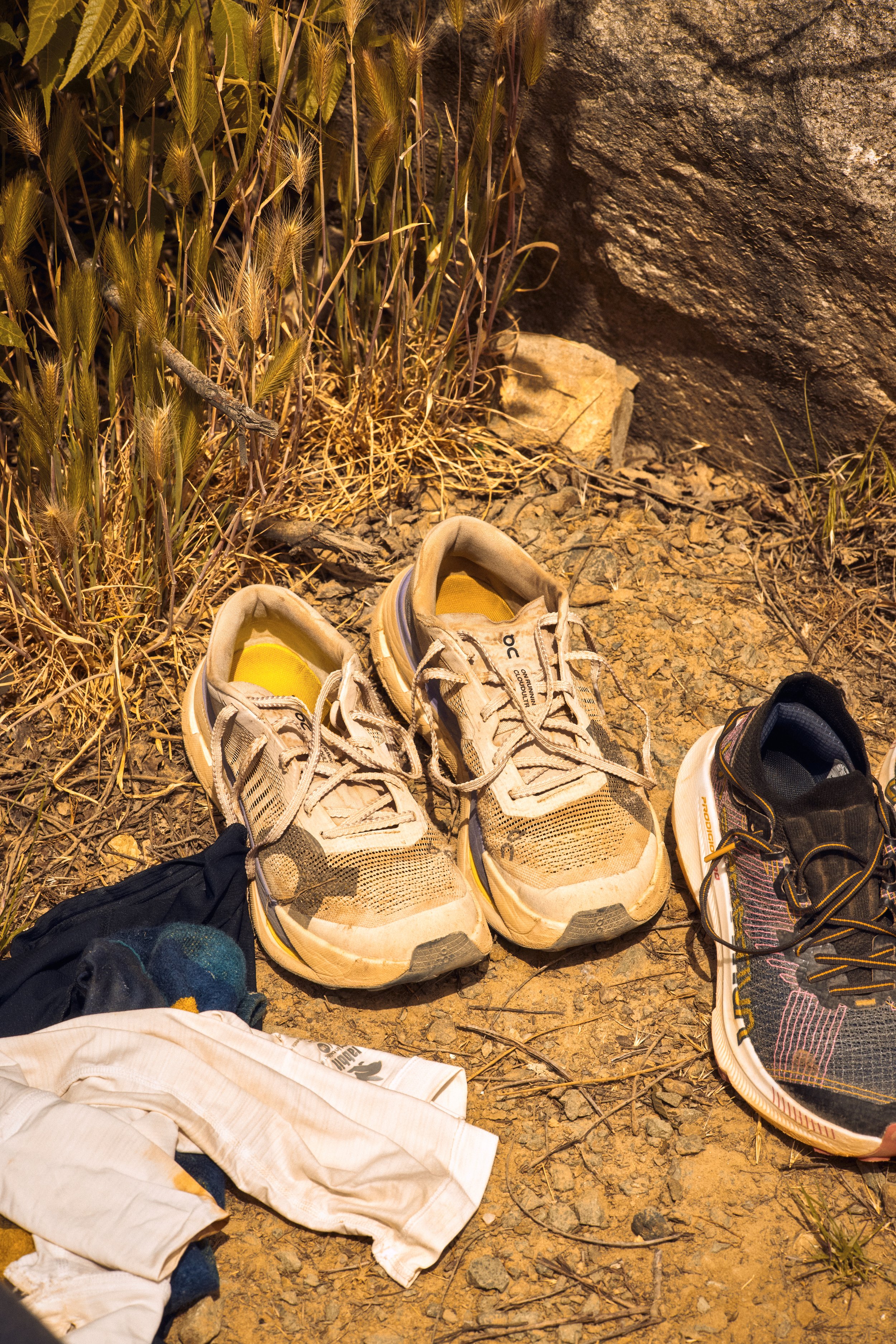 Athletic shoes and clothing placed on dirt ground beside dry grass and a large rock, with a rocky wall in the background.