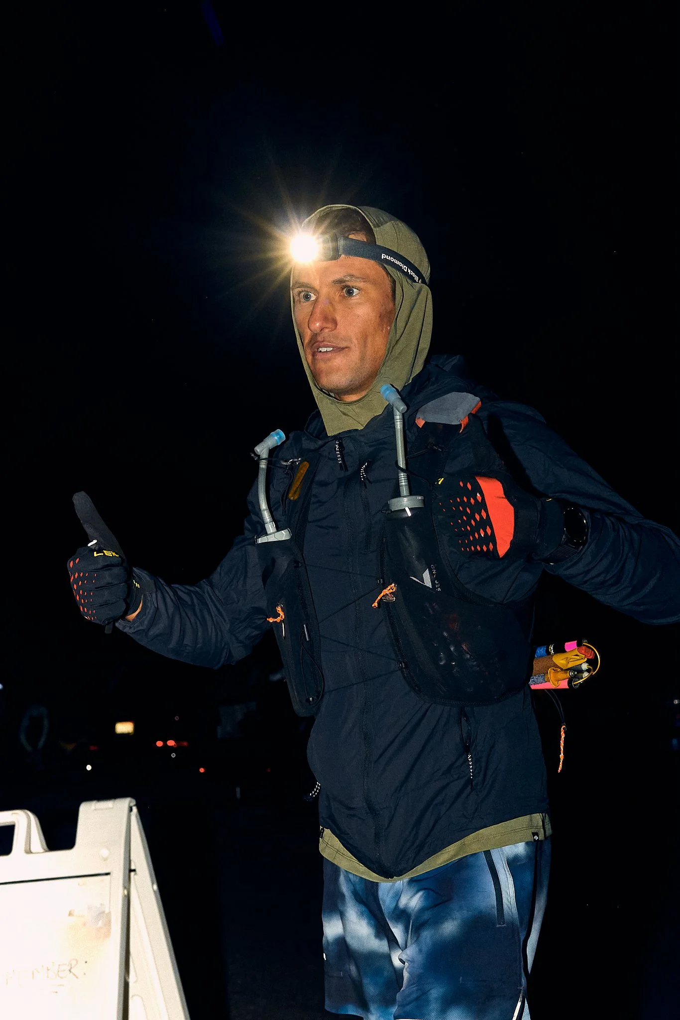 A man wearing outdoor gear and a headlamp giving a thumbs-up at night, standing outdoors with a white sign nearby.
