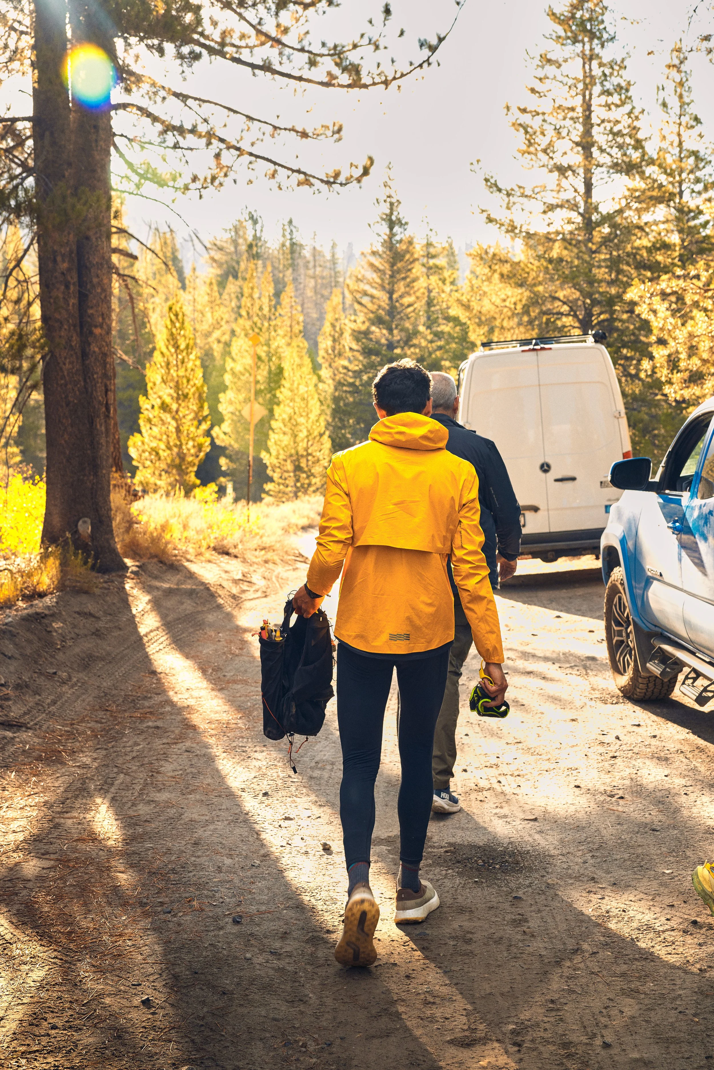 Two people walking on a dirt trail in a forested area during sunset. One person wears a yellow jacket and is carrying a black bag, while the other is dressed in darker clothing. parked cars and a white van are visible on the side of the trail.