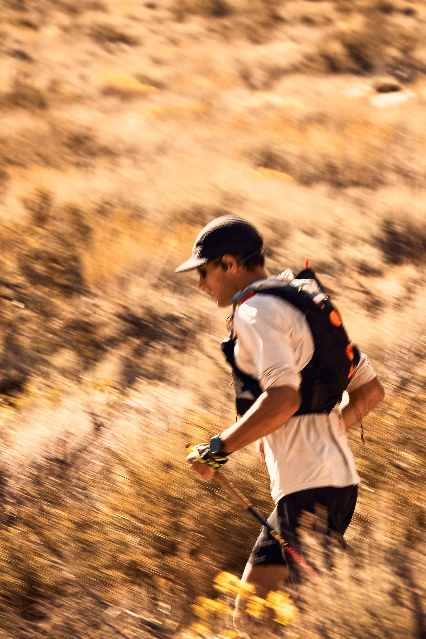 A person running outdoors in a desert-like landscape with blurred background, wearing a white shirt, black shorts, gloves, a cap, and carrying a small backpack.
