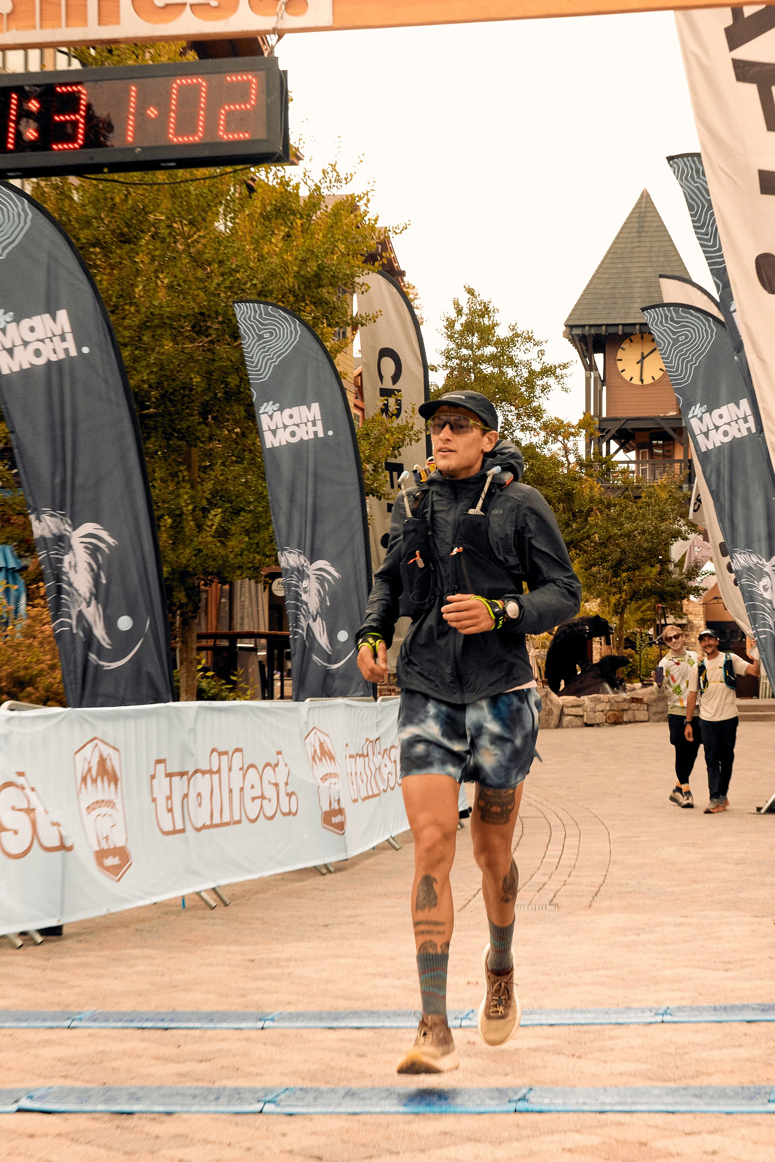 A male runner in black athletic clothing and sunglasses crossing the finish line with a wooden clock tower and trees in the background during a race event.