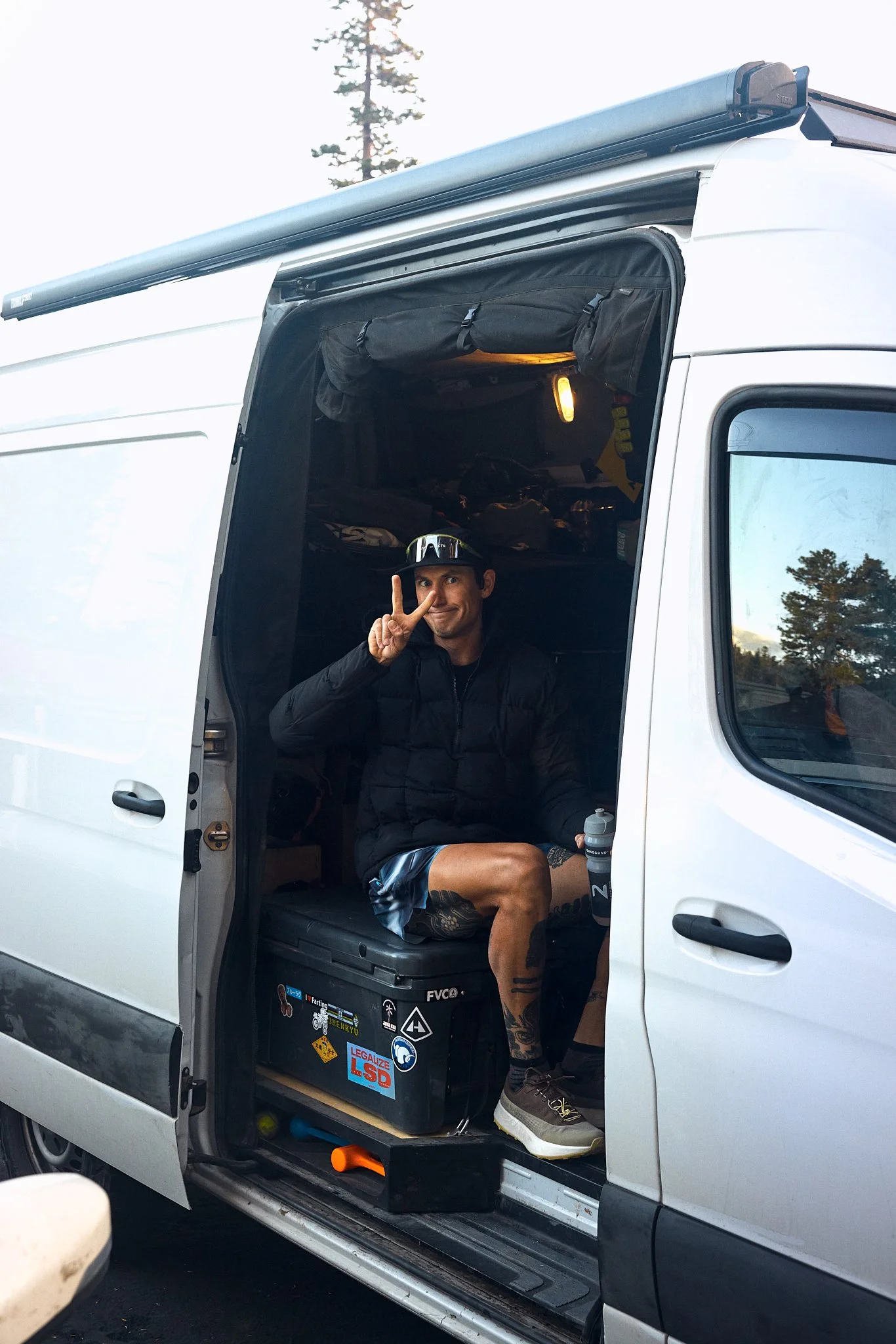 A man sitting inside a white van with the door open, making a peace sign with his right hand. He is wearing a black jacket, shorts, and brown shoes, with tattoos on his left leg. The van's interior has a black container with stickers and a water bott