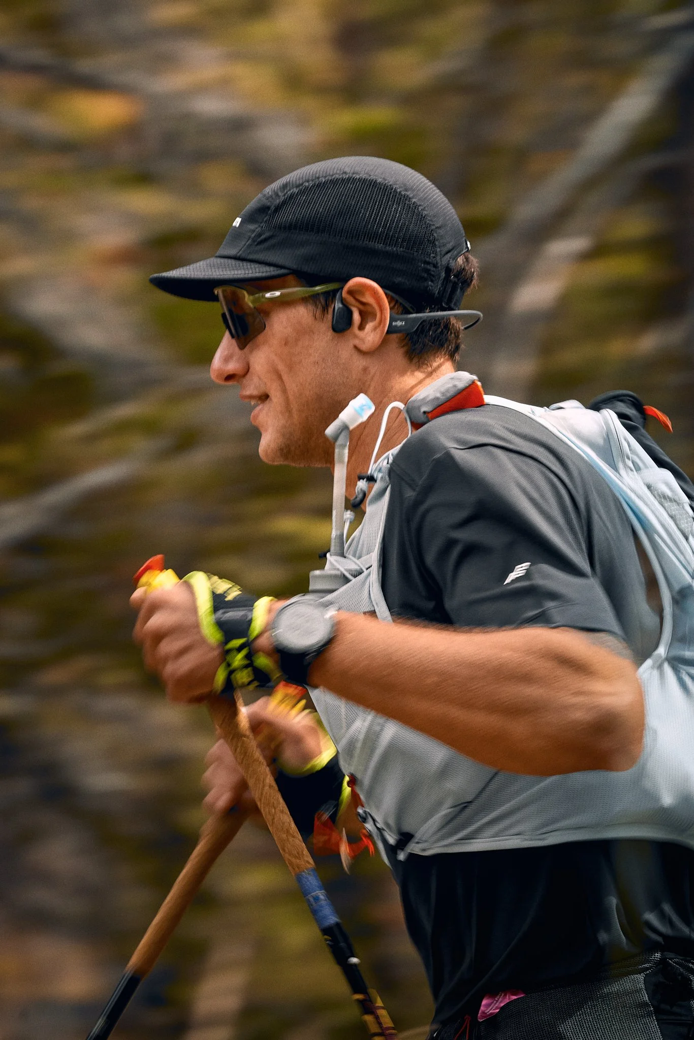 A male trail runner wearing a black cap, sunglasses, and a gray running vest, running outdoors in a natural setting.