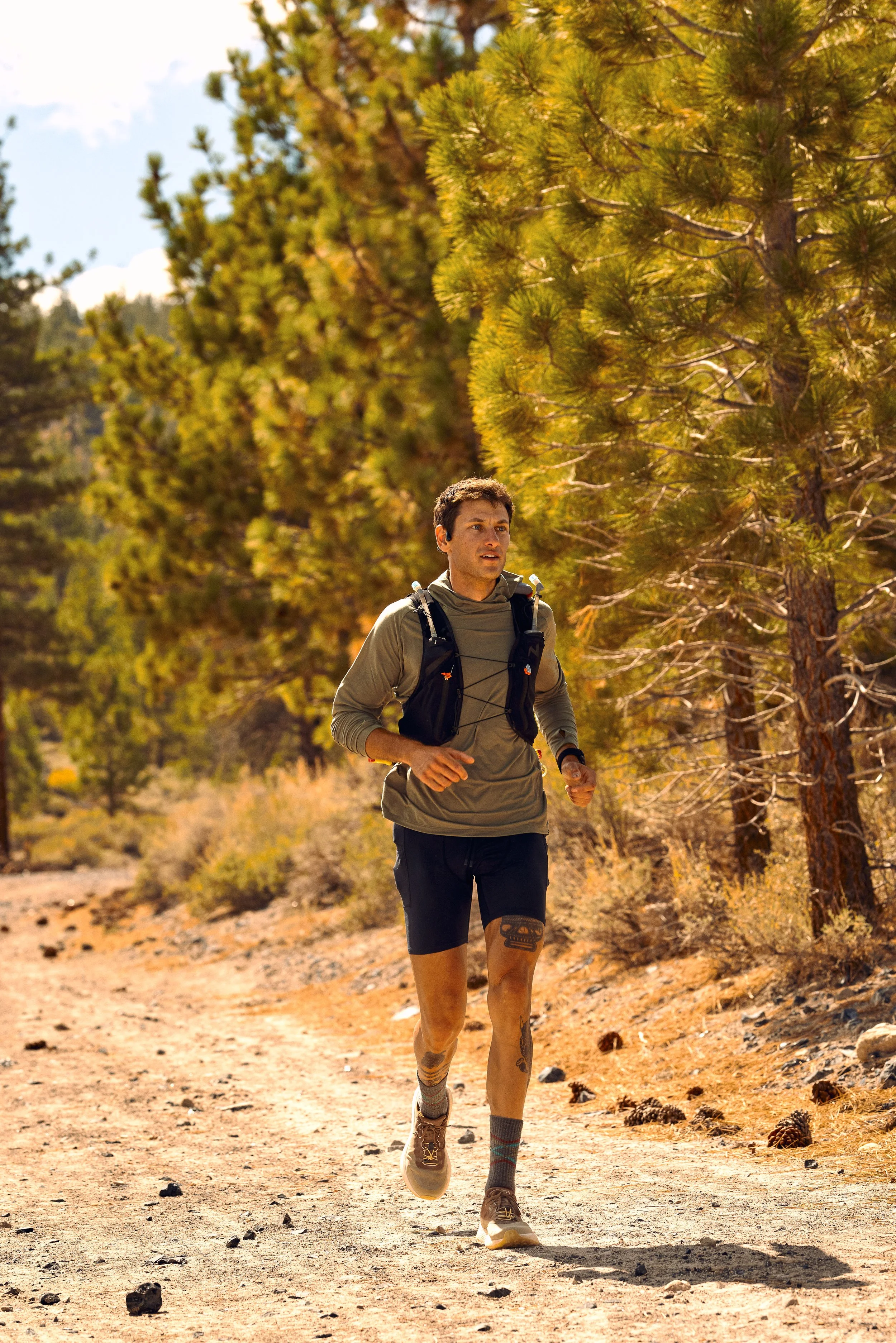 A man running along a dirt trail in a forested area with pine trees under a bright blue sky.