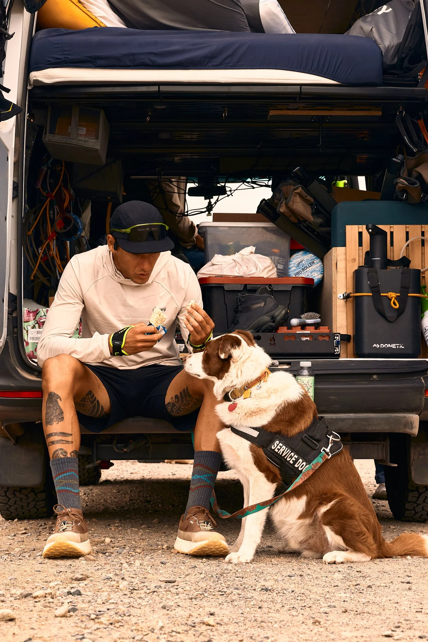A man with tattoos sitting with a service dog in front of an open vehicle packed with outdoor gear and supplies.