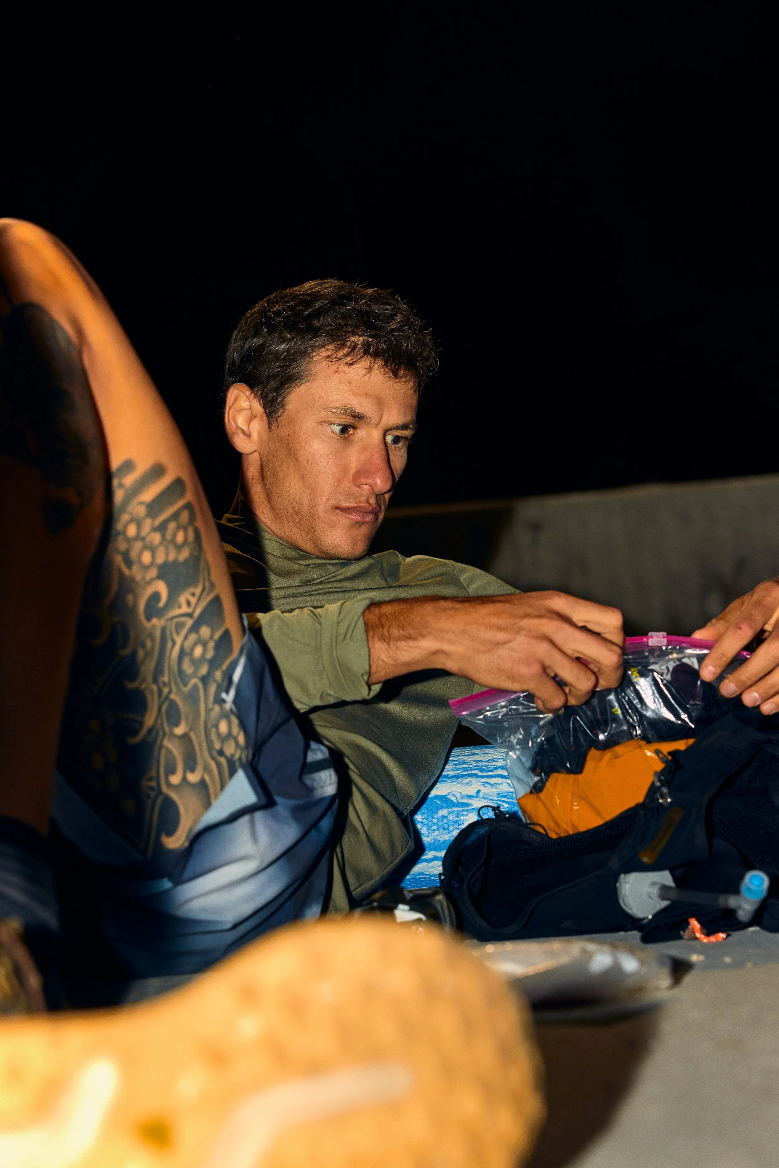 A young man with short dark hair looking at a plastic bag, with a tattooed arm visible in the foreground; other items like orange gear are on the table.