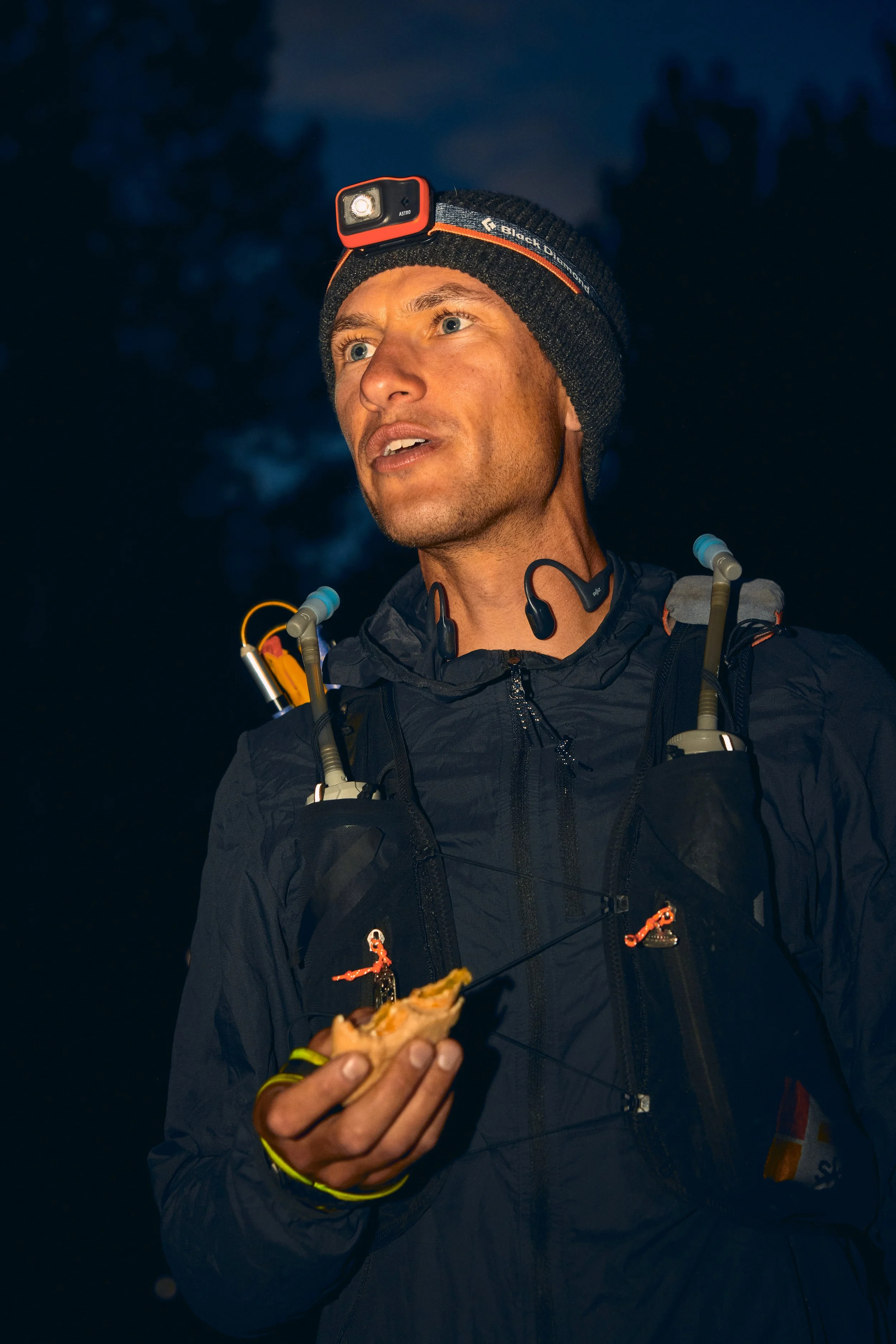 A man outdoors at night wearing a headlamp, a black jacket, and a black beanie hat. He's holding a sandwich or food item in his hand.