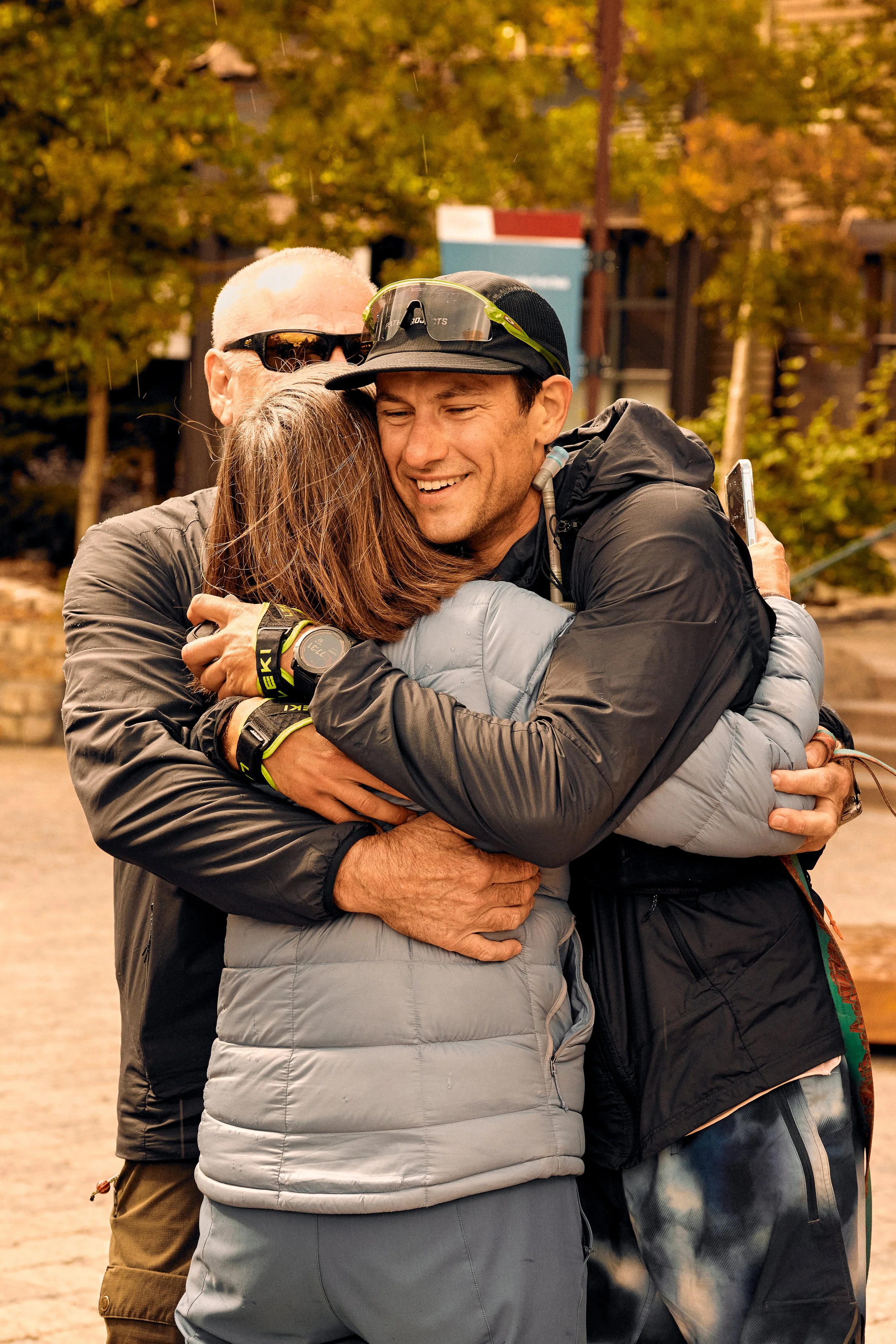 Three people hugging outdoors on a cloudy day, with fall foliage in the background.