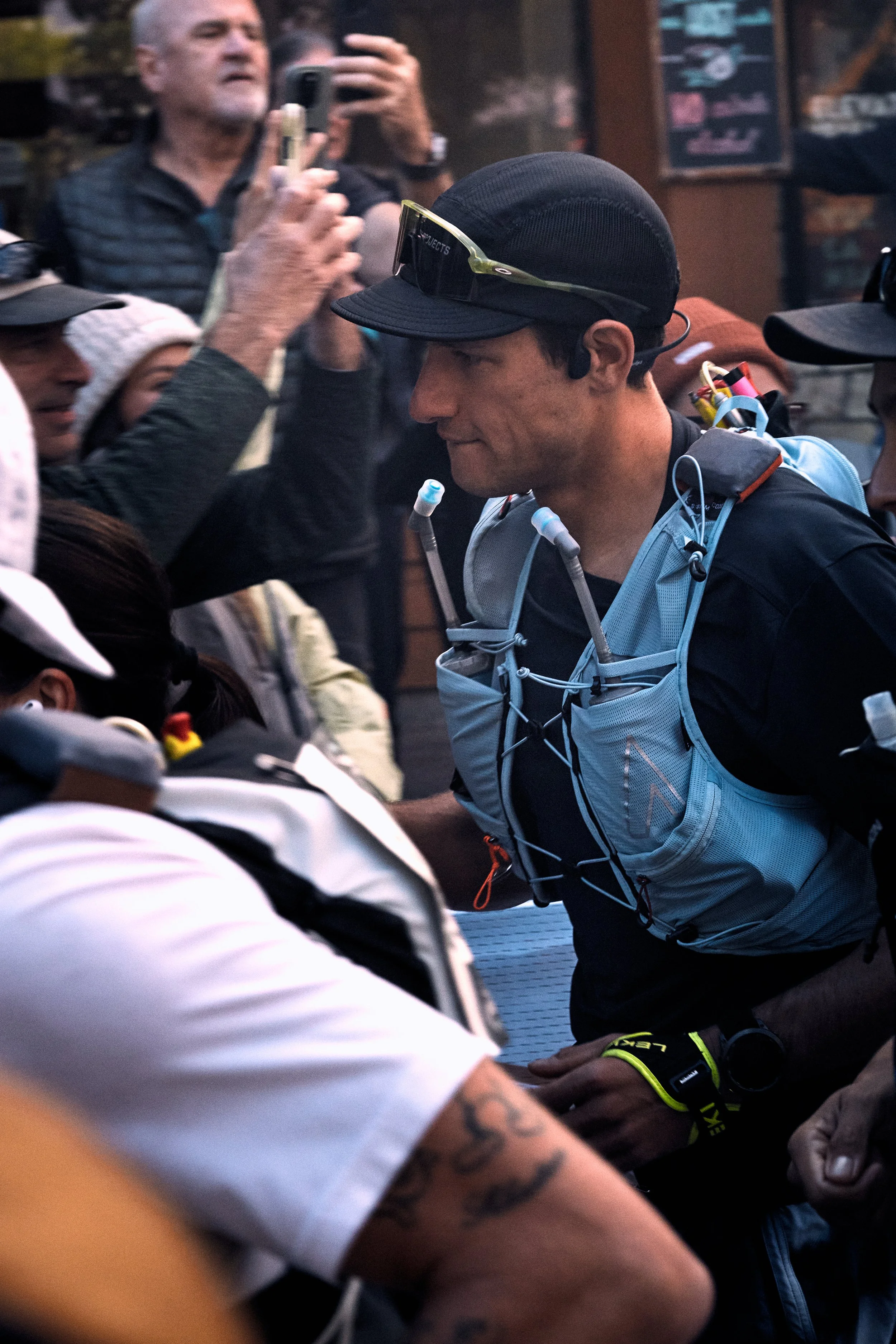 Close-up of a man wearing a black cap, sunglasses on his forehead, with a hydration pack, in a crowded outdoor setting. Other people around him are taking photos and appear to be in a race or event.