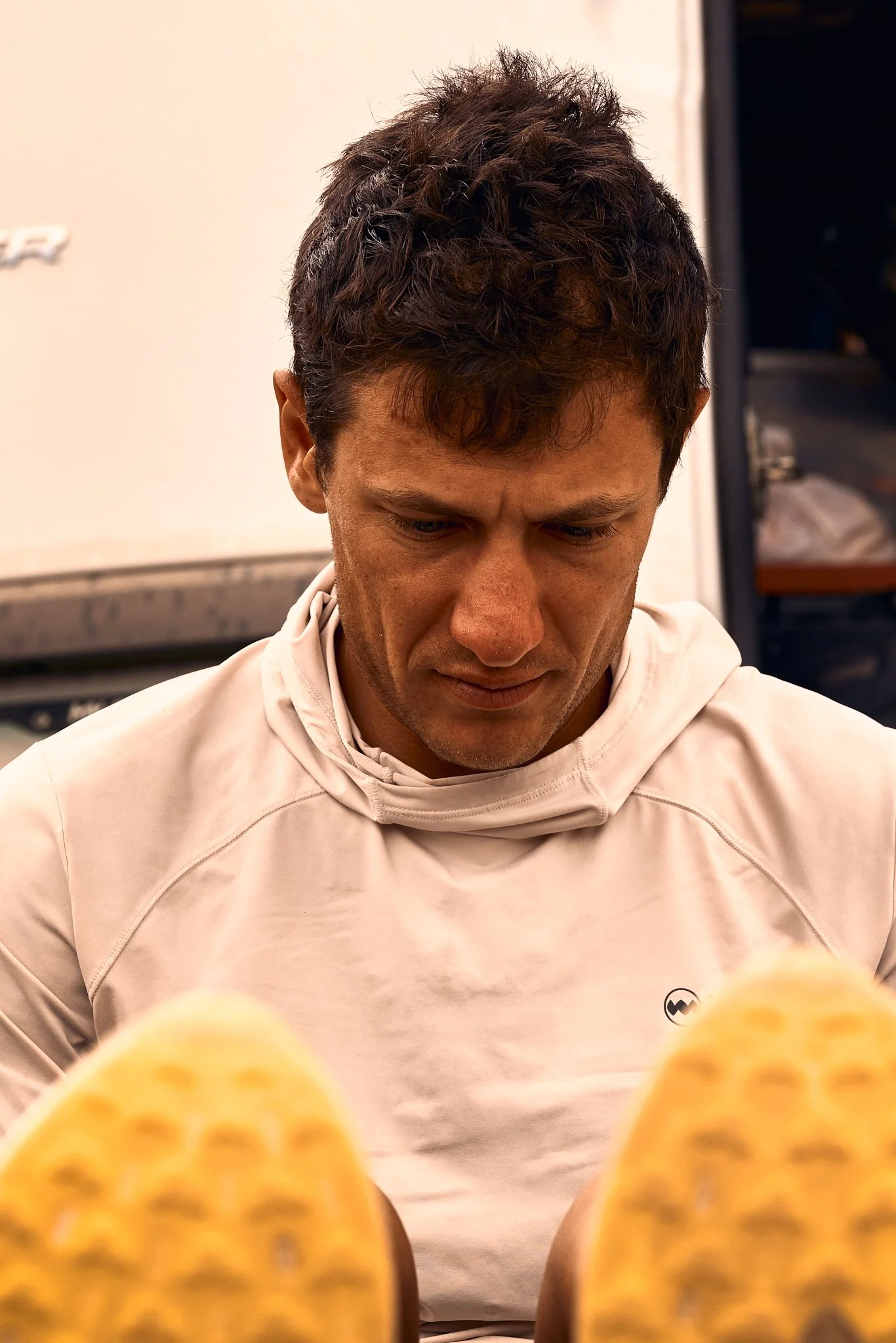 A man with short curly brown hair and light skin is looking down, wearing a light beige hoodie with a small black logo on the chest. The background shows a wall and a partially open door with a dark interior.