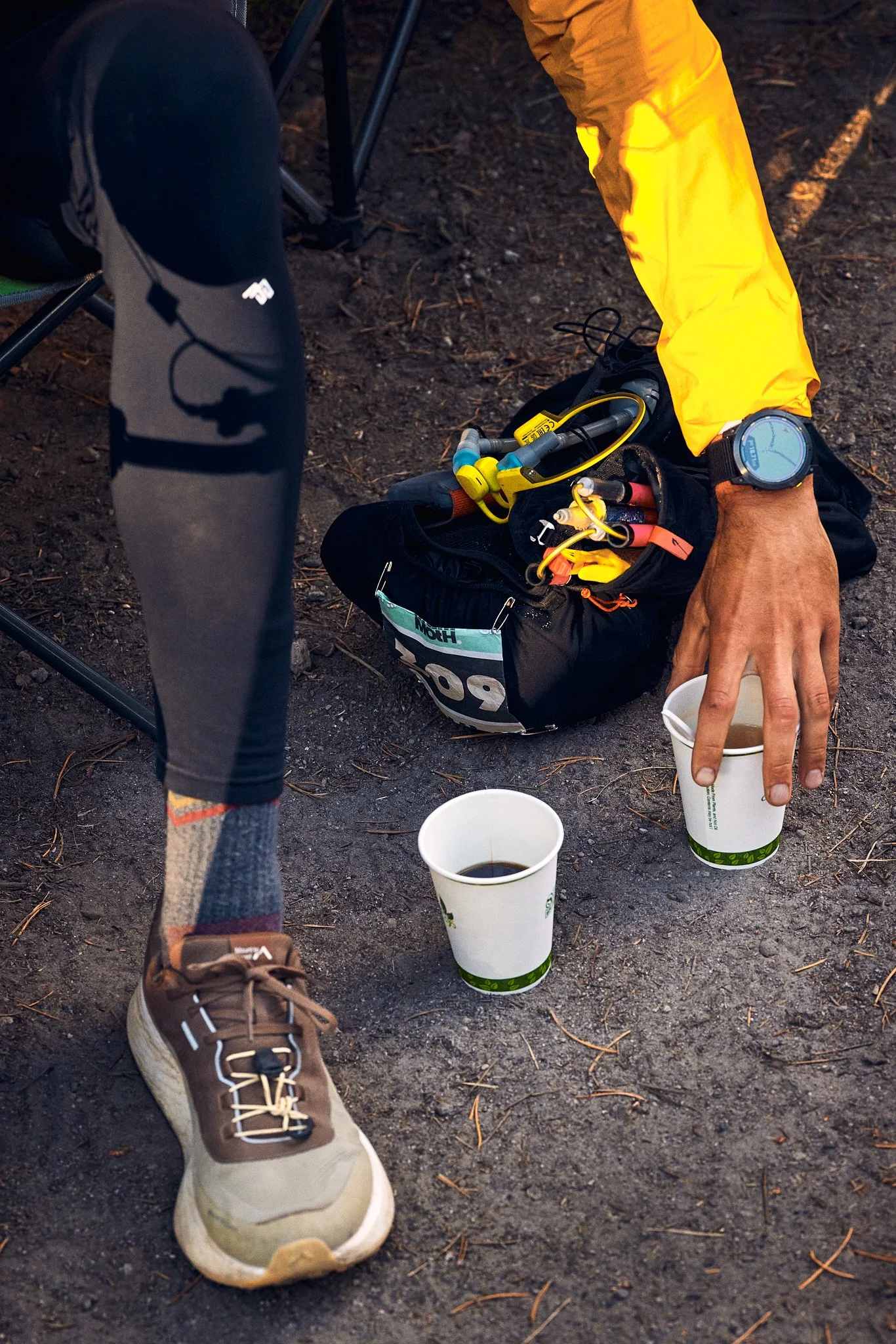 Outdoor scene with a person wearing hiking shoes, yellow waterproof jacket, and a watch, seated on the ground near a backpack containing climbing or hiking gear. Two cups of coffee or tea are on the ground nearby, with a person's hand reaching for on