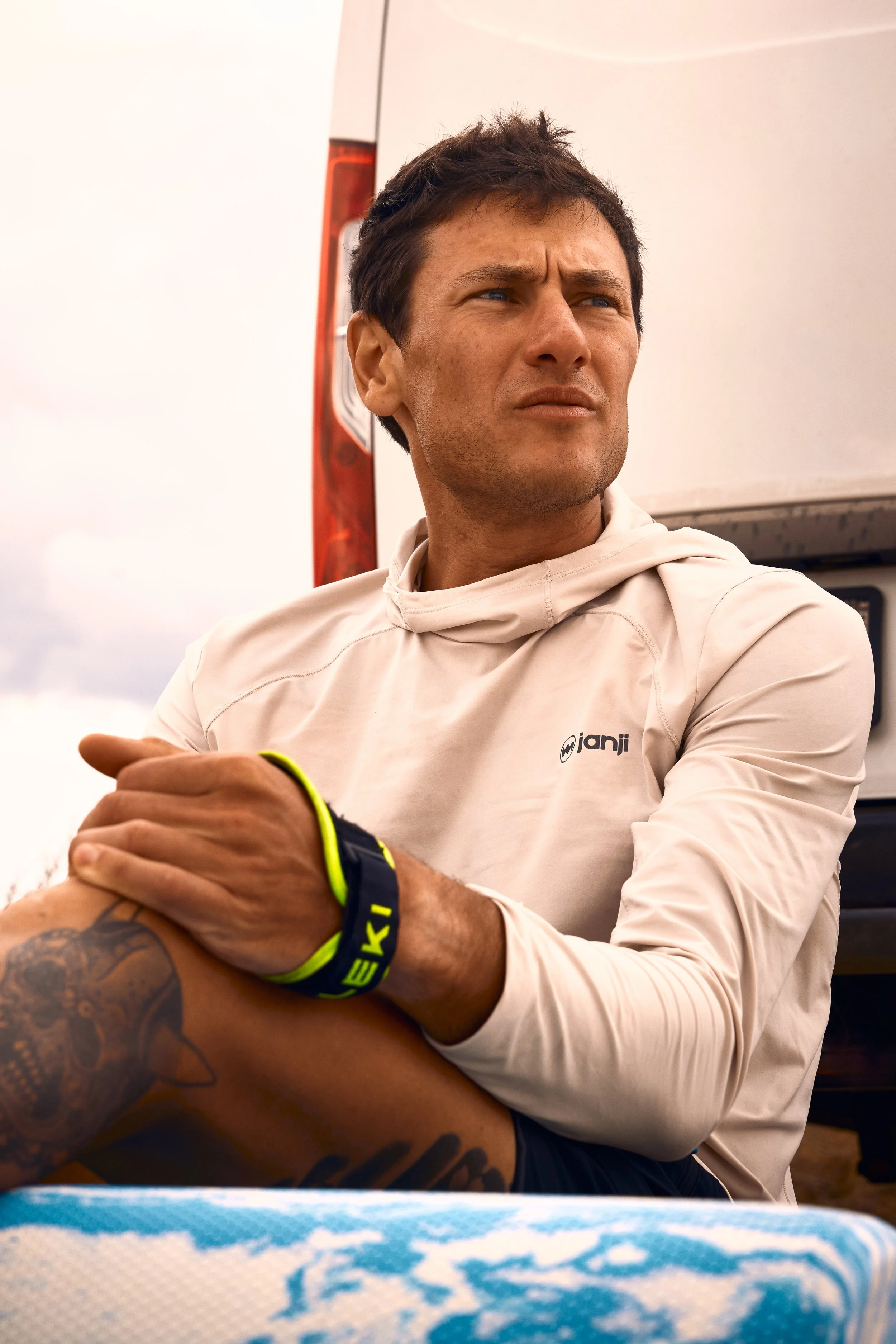 A man with short, dark hair and tan skin looking to his left, wearing a cream-colored hoodie with a logo, with a serious expression. He has a black wristband with yellow lettering and a tattoo on his left arm. The background includes a truck or large