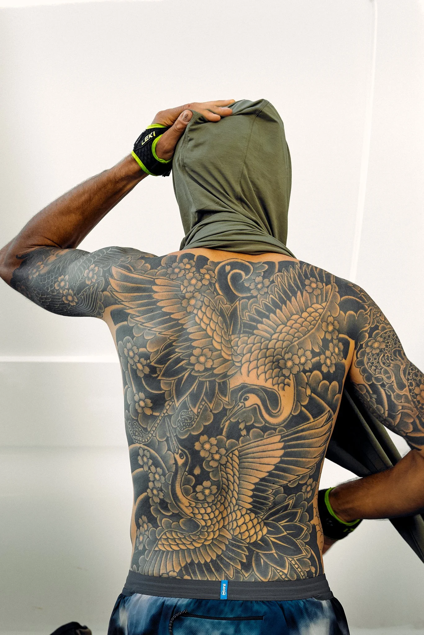 A shirtless man with a large detailed Japanese dragon tattoo on his chest and arm, wearing a green hoodie over his head, a black wristband, and black and blue athletic shorts, standing against a plain white wall.