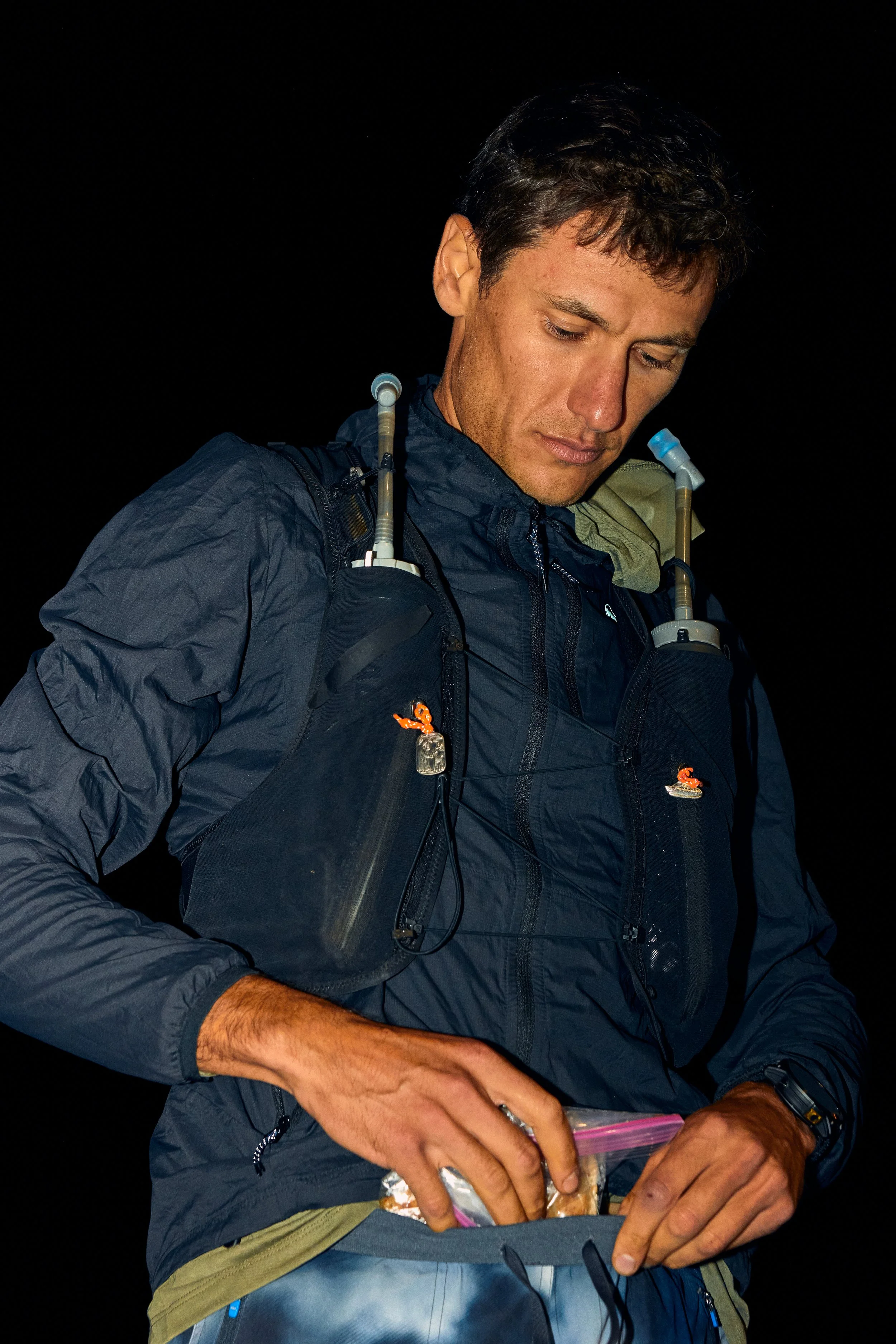 A man with short dark hair wearing a dark blue jacket with a tan and black backpack, looking down at a small plastic bag, in a dark environment.