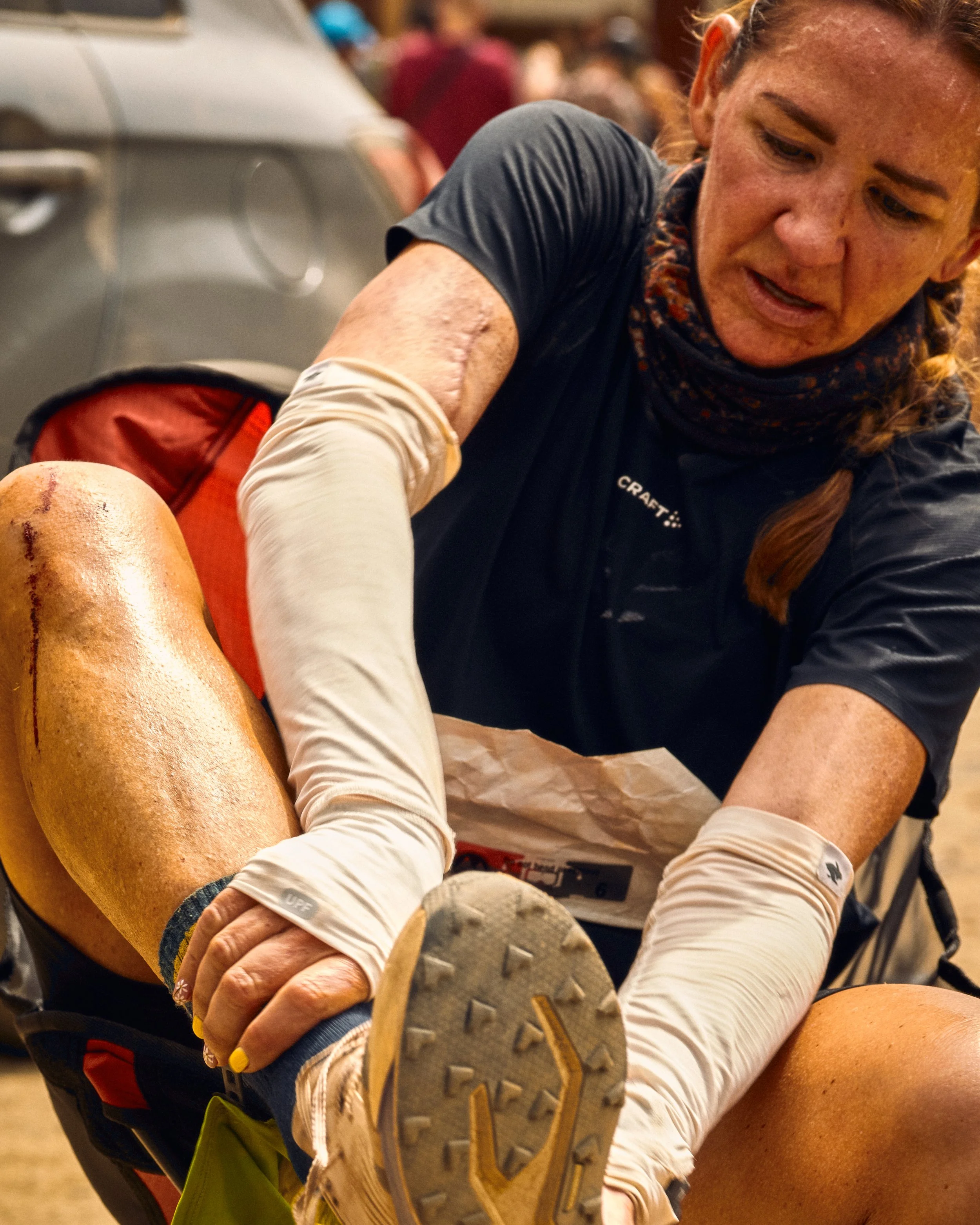 Close-up of a woman with a curly ponytail, wearing athletic clothing and gloves, sitting with her foot extended and resting on her knee, in a crowded indoor setting.