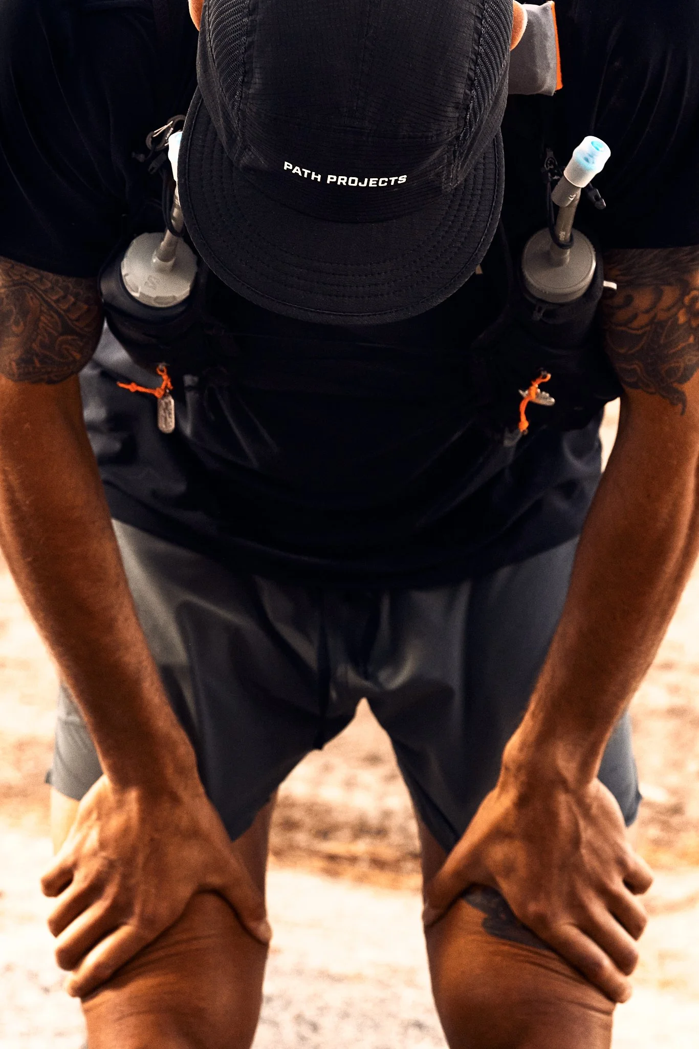 A person with tattoos on their arms wearing a black baseball cap with white text that reads 'PATH PROJECTS', a black backpack, and dark shorts, sitting with hands on knees. The image is taken from above, focusing on the person's torso and arms.