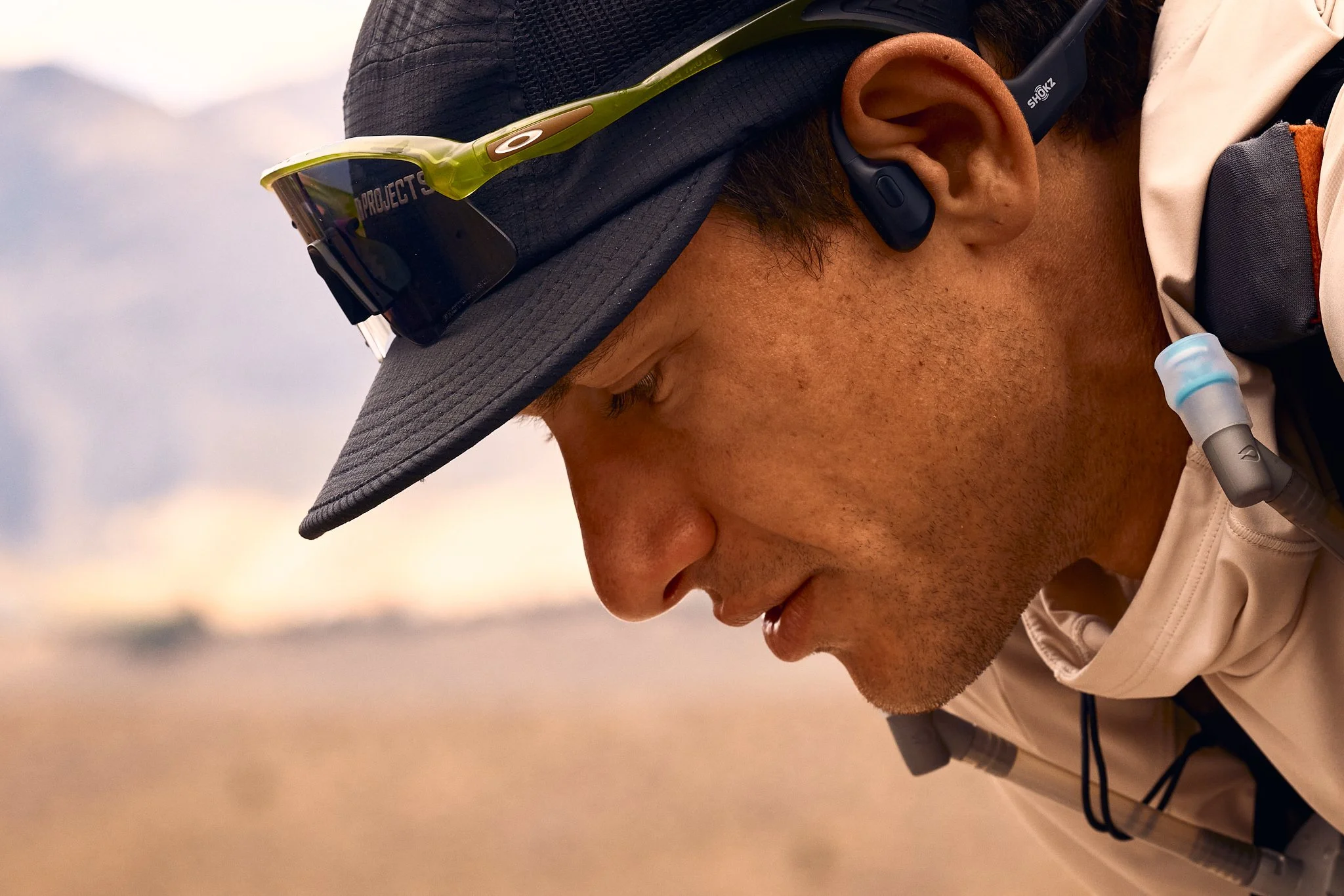 Close-up of a person wearing a gray cap, sunglasses, and a headset with a microphone, outdoors during sunset.