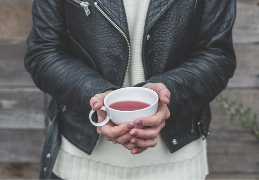 Person holding a white cup with tea, wearing a black leather jacket and white sweater, standing in front of a wooden background.