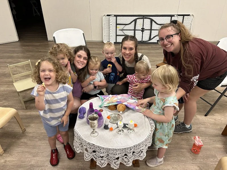 Children and an adult gathered around a small table with a white lace tablecloth, celebrating a birthday with a cake, snacks, and decorations in a room with wooden flooring and chairs.