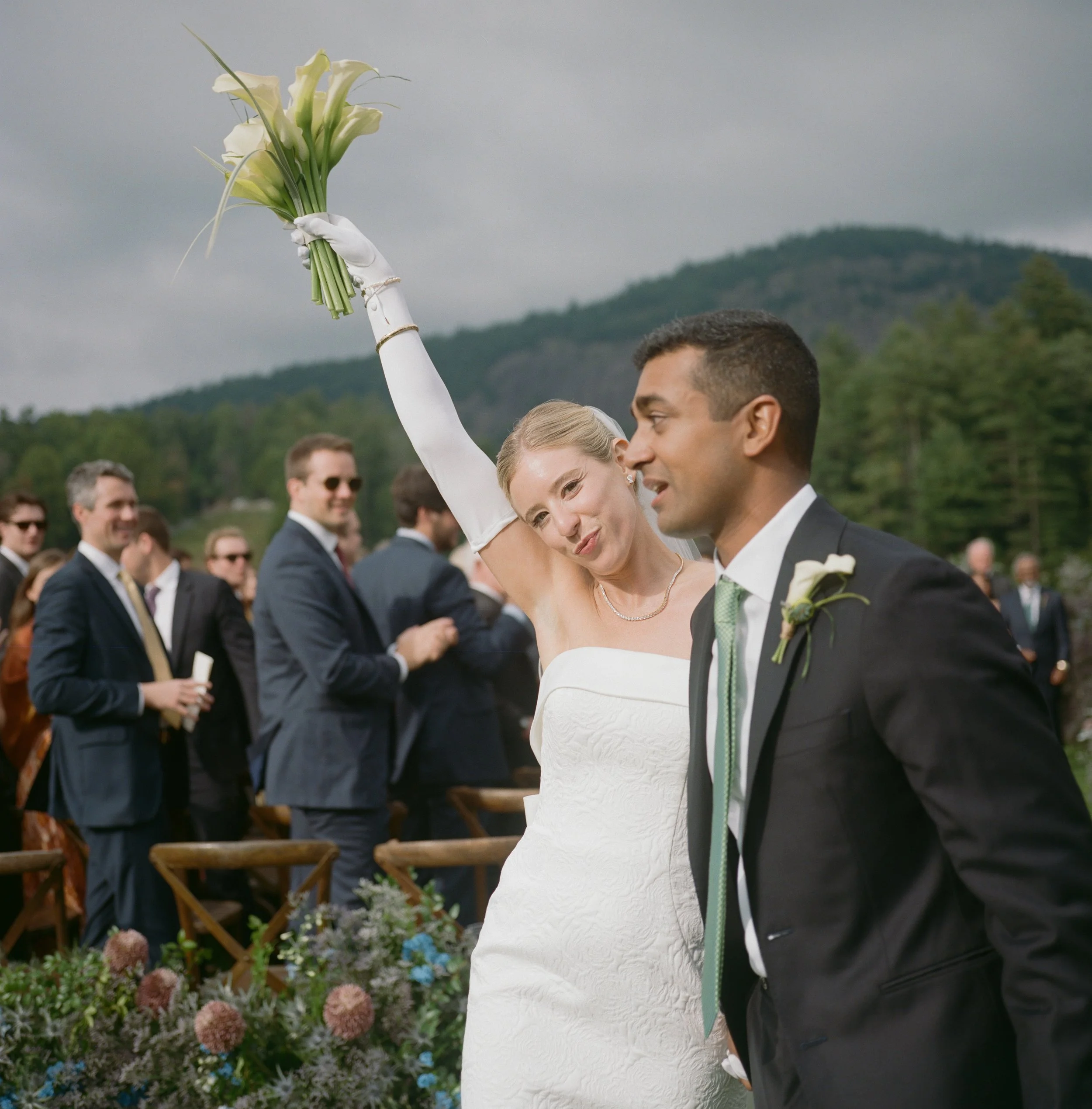 Bride holding a bouquet of white flowers and celebrating with guests outdoors at a wedding ceremony.