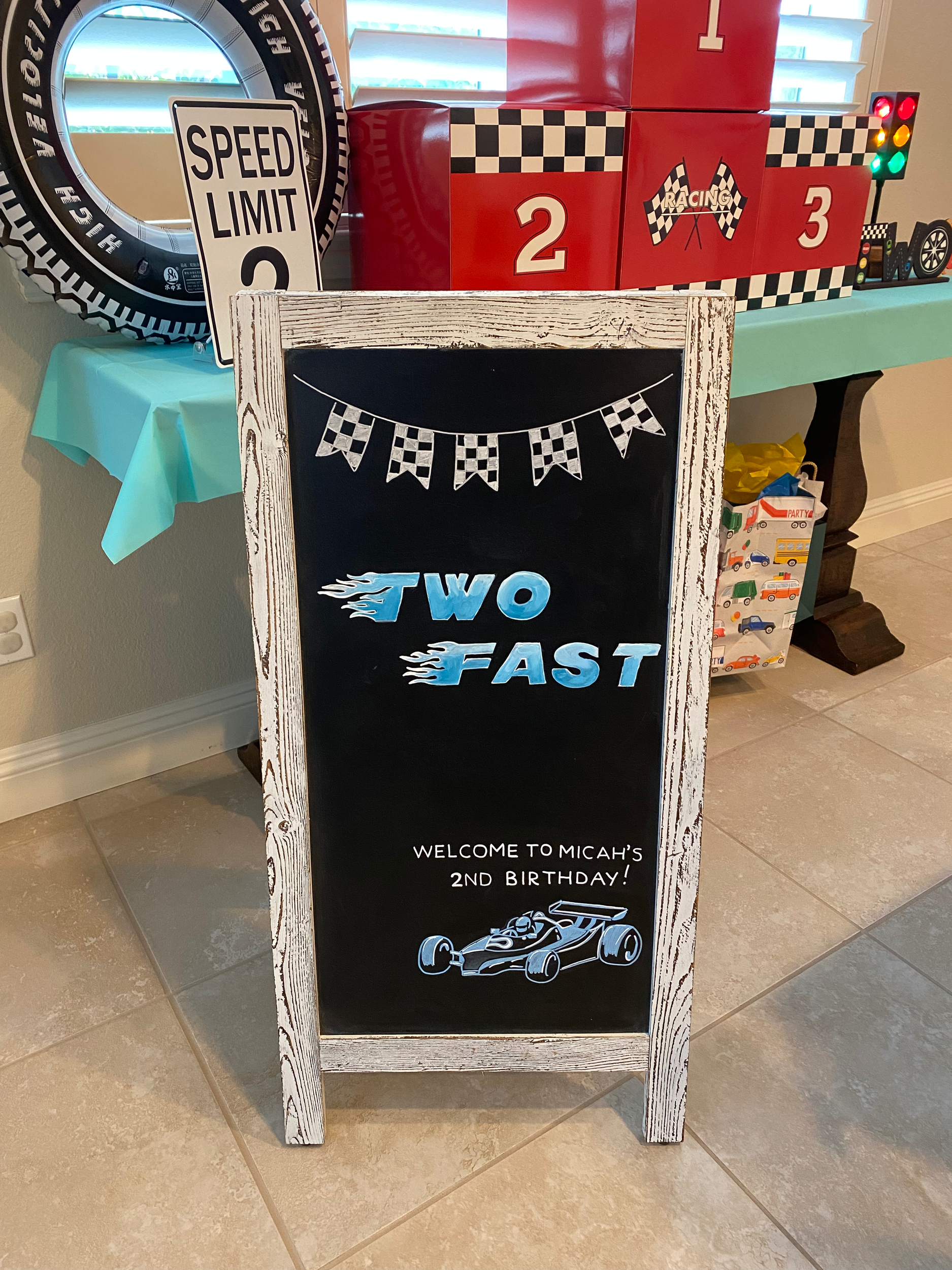 Hand lettered and illustrated chalkboard that says "Two Fast" for a 2 year old birthday party. In the bottom right corner is a hand illustrated race car and at the top are hanging black and white checkered race track flags.