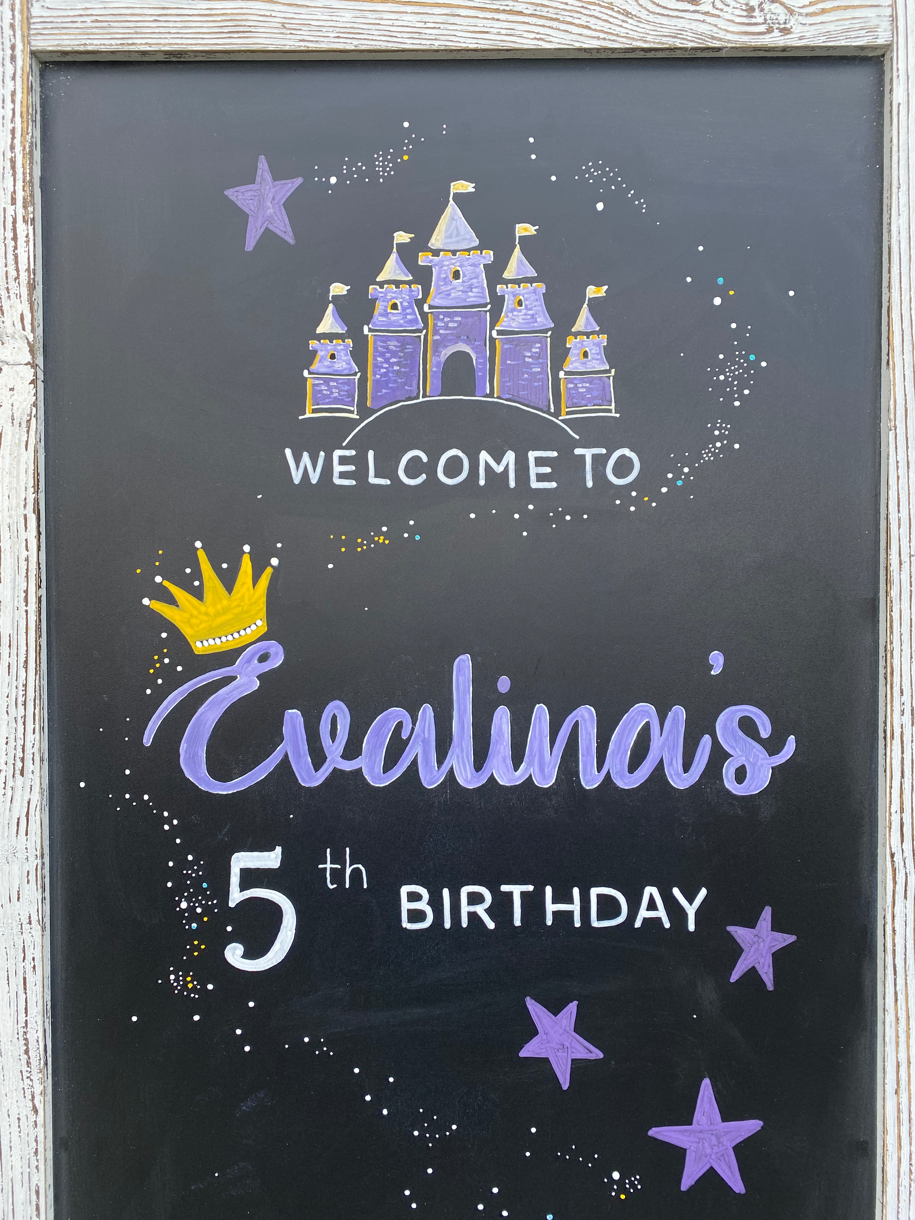 A close up image of a hand lettered and hand illustrated chalkboard for a 5 year old princess birthday. At the top is a purple castle with a stream of shooting stars. At the bottom is a blue, purple, and yellow rainbow with whimsical clouds. It says 