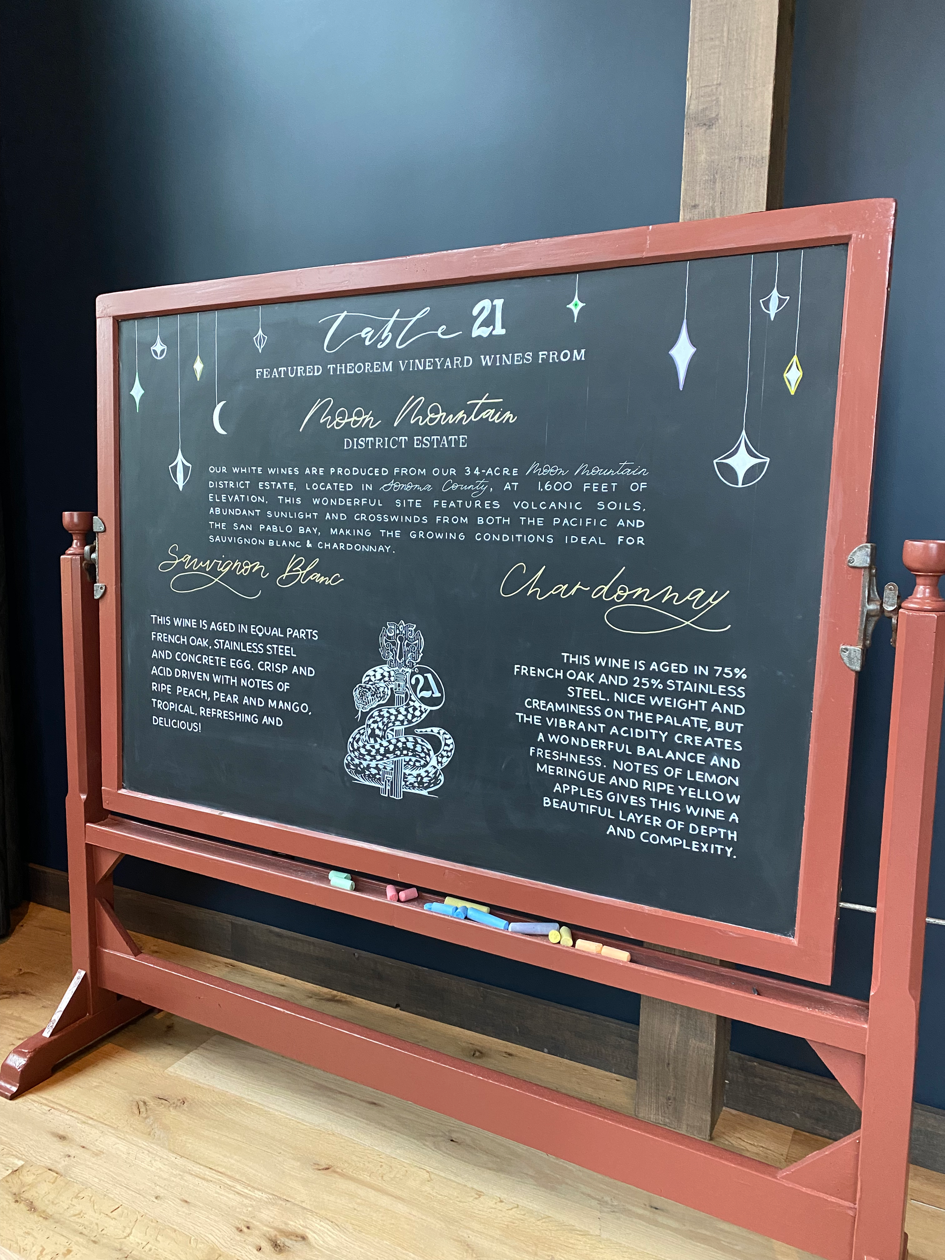 A large display chalkboard hand lettered for Theorem Vineyards in Napa Valley. There is a large description of two wines, Sauvignon Blanc and Chardonnay, from Moon Mountain district, decorated with celestial designs appearing to hand down from the to