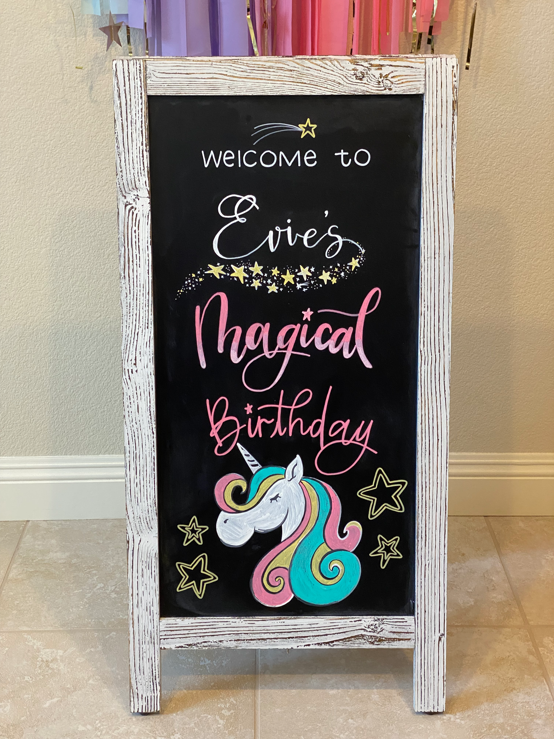 A birthday chalkboard hand lettered and hand illustrated to say "welcome to Evie's Magical Birthday" decorated with bright yellow stars, pink and white writing, and a unicorn with a pink and blue swirly mane.