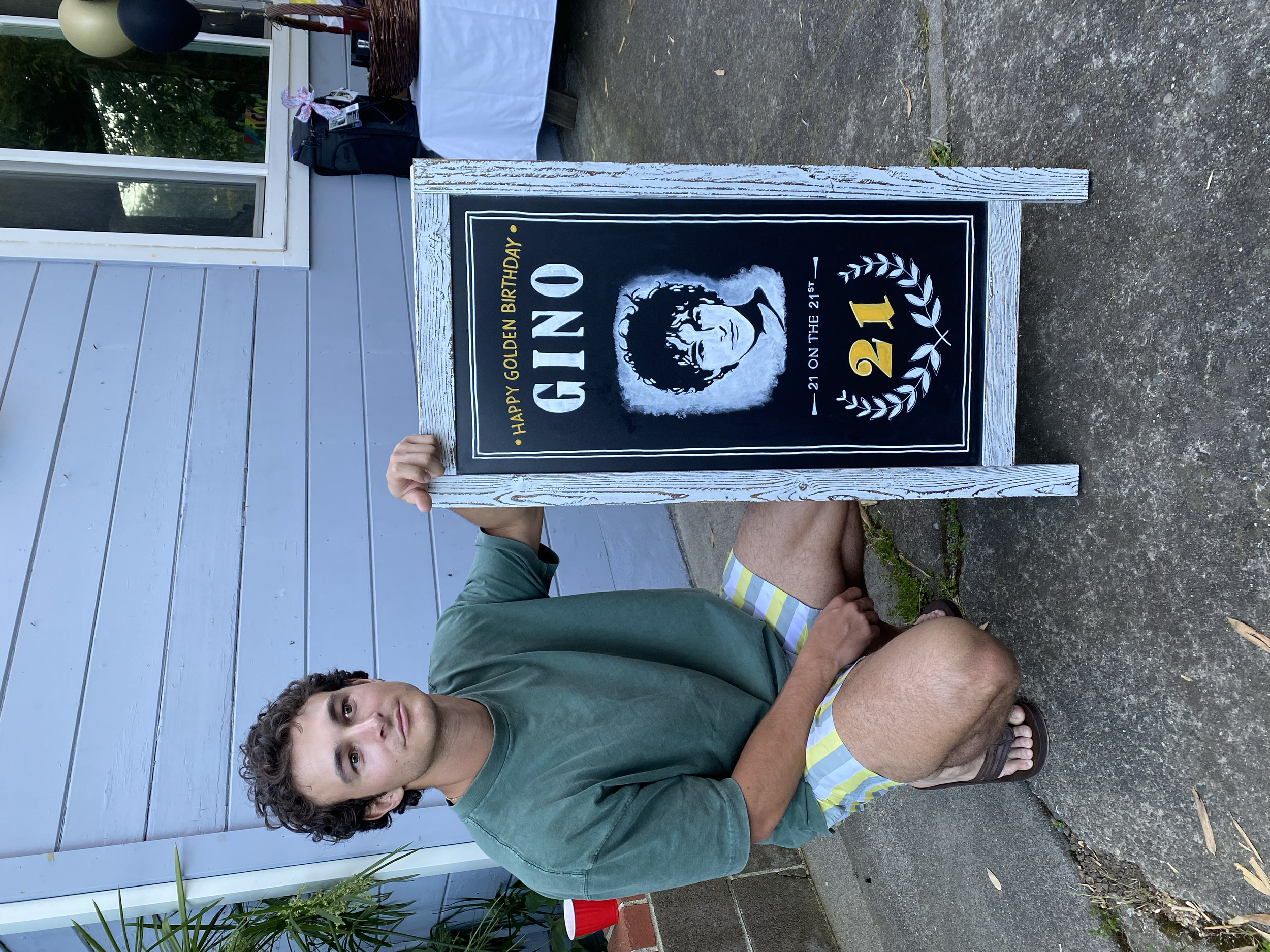 Hand lettered and illustrated 21st birthday chalkboard that says Happy Golden Birthday Gino, pictured with Gino. 21st on the 21st, with a chalkboard illustration portrait of the birthday boy's face. Below are large yellow numbers for 21