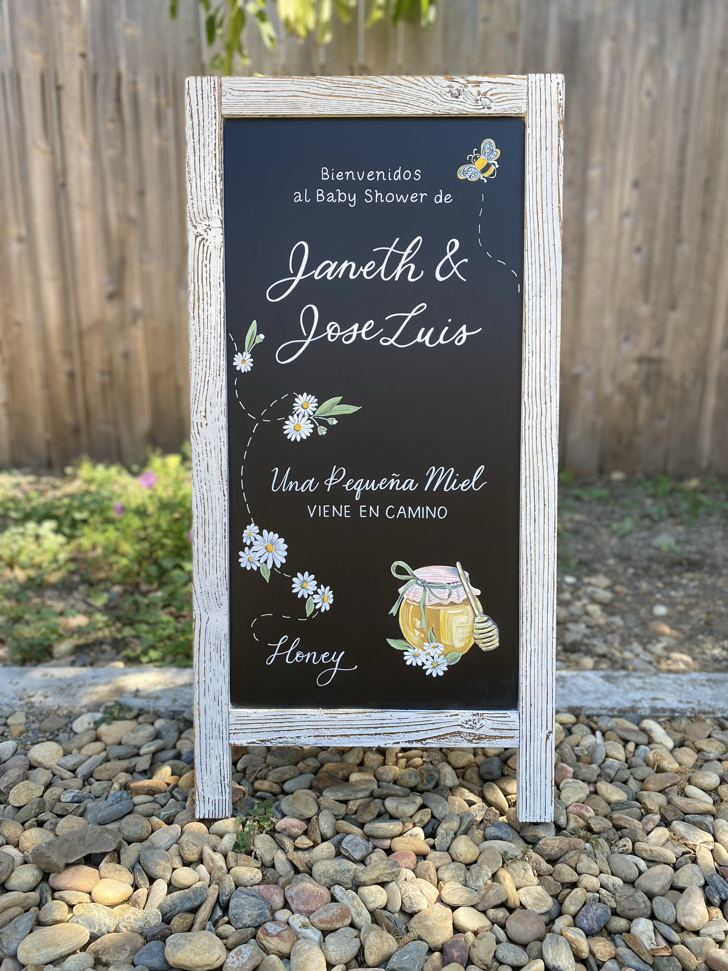 A hand lettered and illustrated baby shower chalkboard, decorated with white daisies, a bee, and a honey pot. 