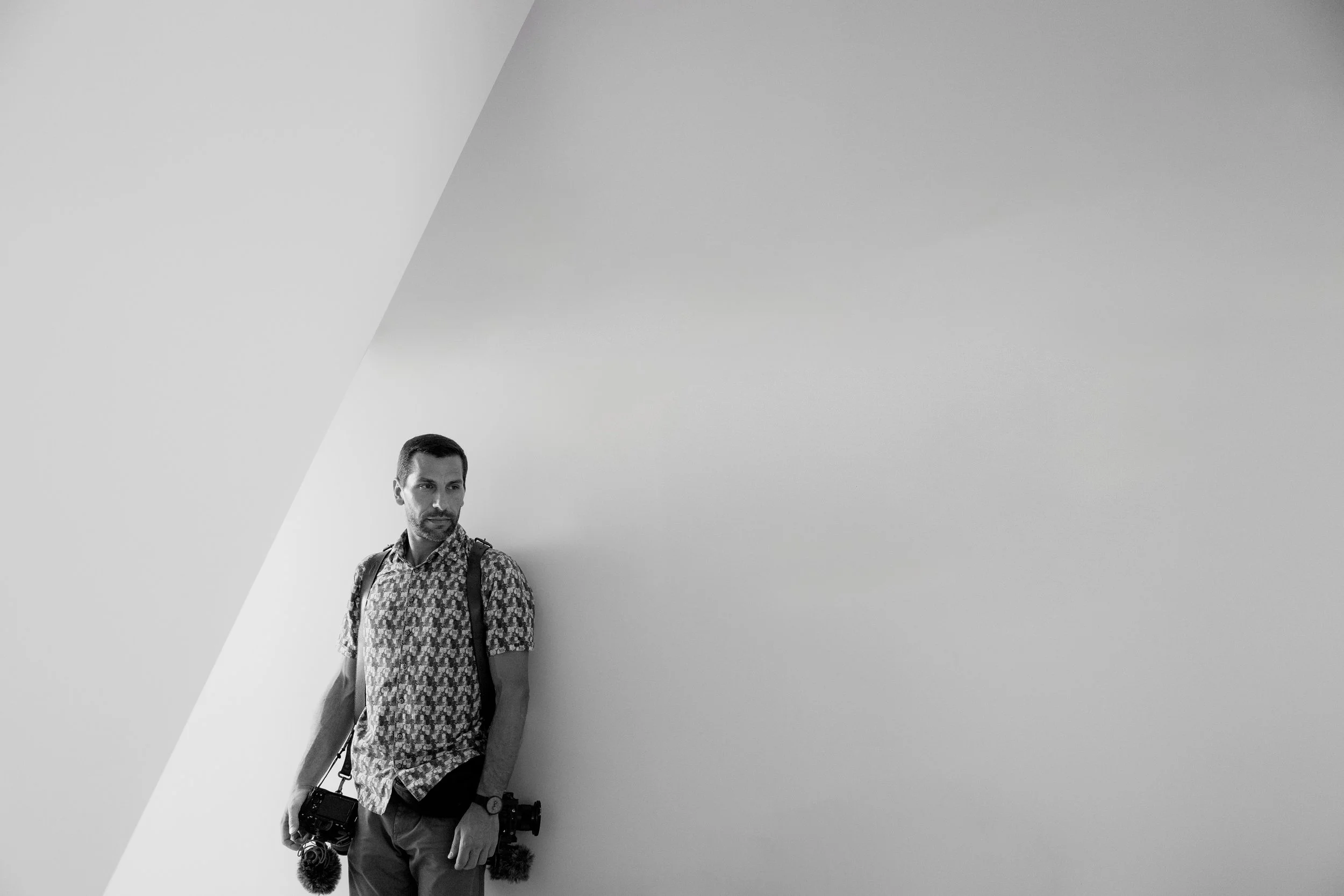 A wedding videographer, holding cameras, stands against a plain wall with a large diagonal shadow, looking to couple